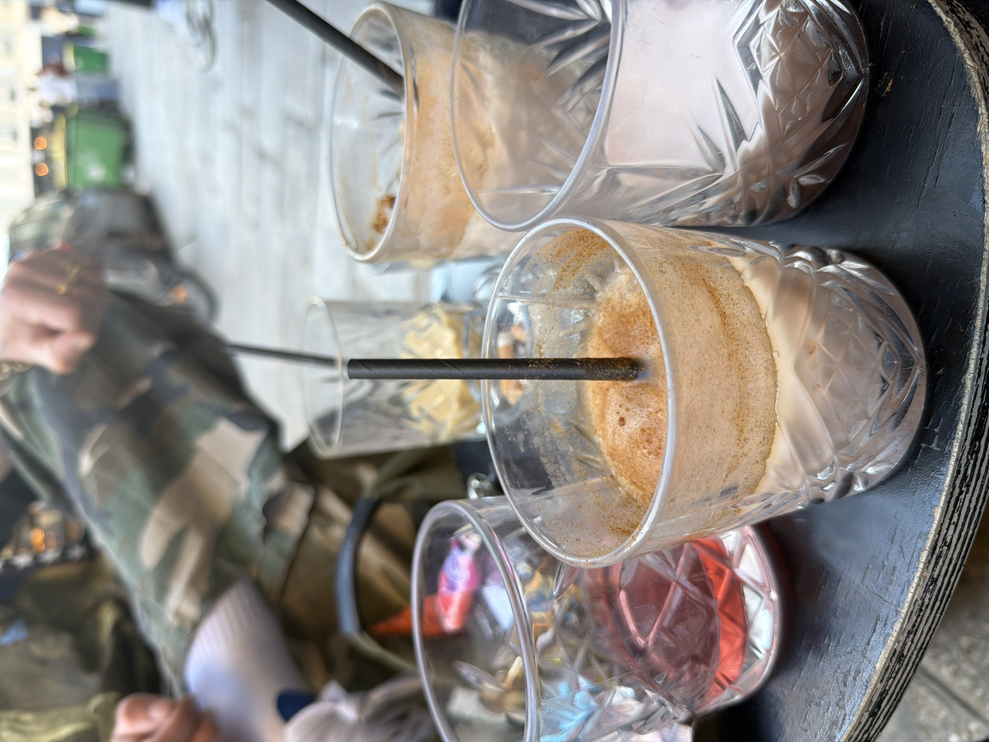 The image presents a close-up view of a round, black table on which four ornate, clear glasses are arranged. Each glass contains varying amounts of liquid, with two showing remnants of a beige, coffee-like beverage, one filled with a pink liquid, and another appearing largely empty. Each is equipped with a black straw. To the left of the frame, a person wearing camouflage pants and white socks is visible, their hand resting near the edge of the table.
The setting appears to be an outdoor café, judging by the presence of a blurred background that suggests a street scene with greenery and other structures. It's plausible the location is Paris, France, given the style and atmosphere commonly associated with Parisian cafes. The glasses look relatively empty suggesting that the patrons are finished with their beverages.