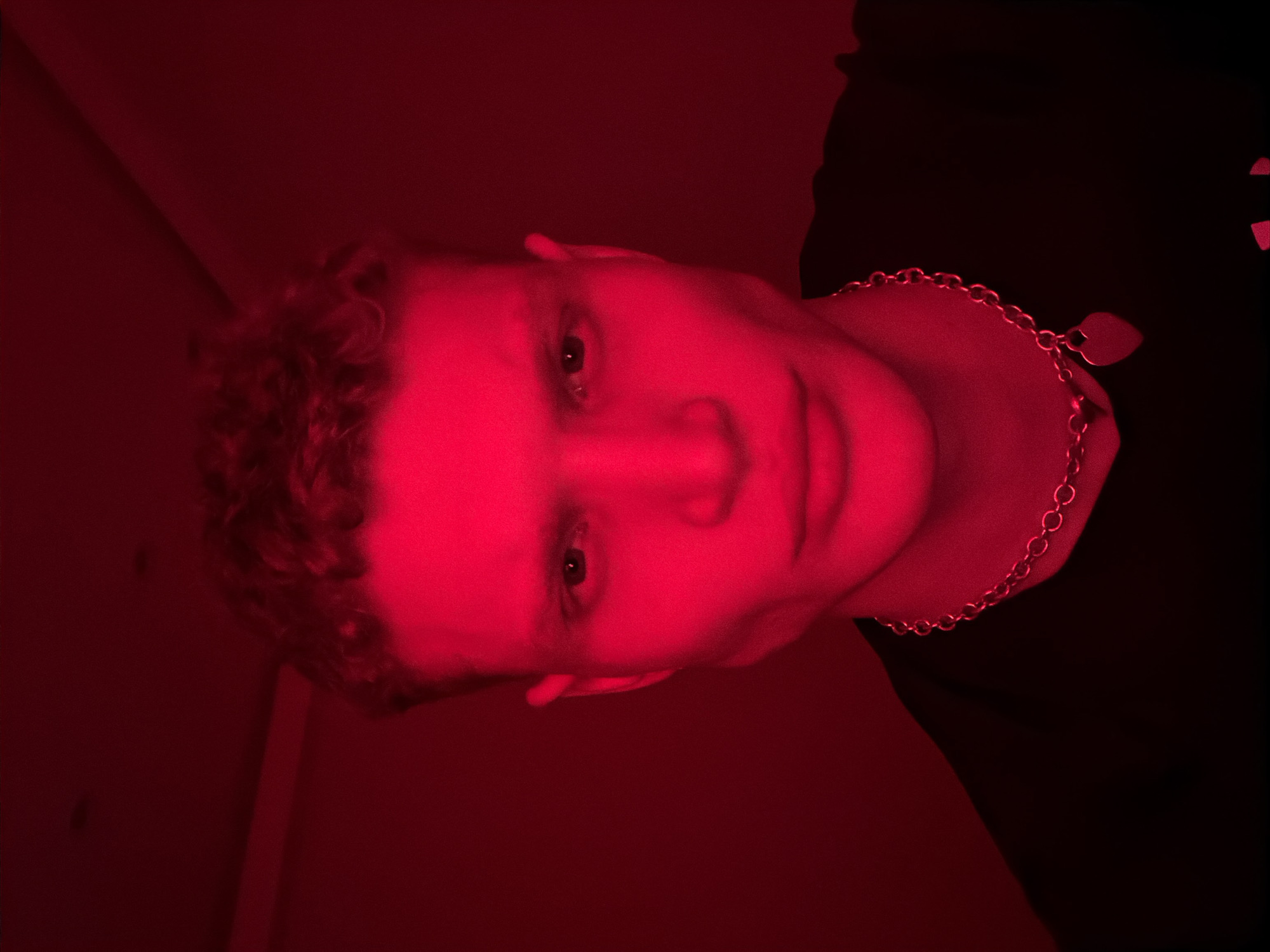 The image displays a close-up portrait of a single individual, appearing male, illuminated entirely by intense red artificial light. The person has short, curly hair and prominent facial features, including a defined jawline and cheekbones. Their eyes are dark, open, and directed straight towards the viewer, with a neutral expression. They wear a dark, possibly black, crew-neck garment and a silver-toned chain necklace with a heart-shaped pendant positioned centrally on their chest. The background is uniformly dark red, indicating an indoor setting with plain walls, possibly a corner visible in the upper left, but no specific room details or external elements are discernible. No visual indicators within the image allow for the determination of a specific location in the city of London, United Kingdom, or any other geographical area.