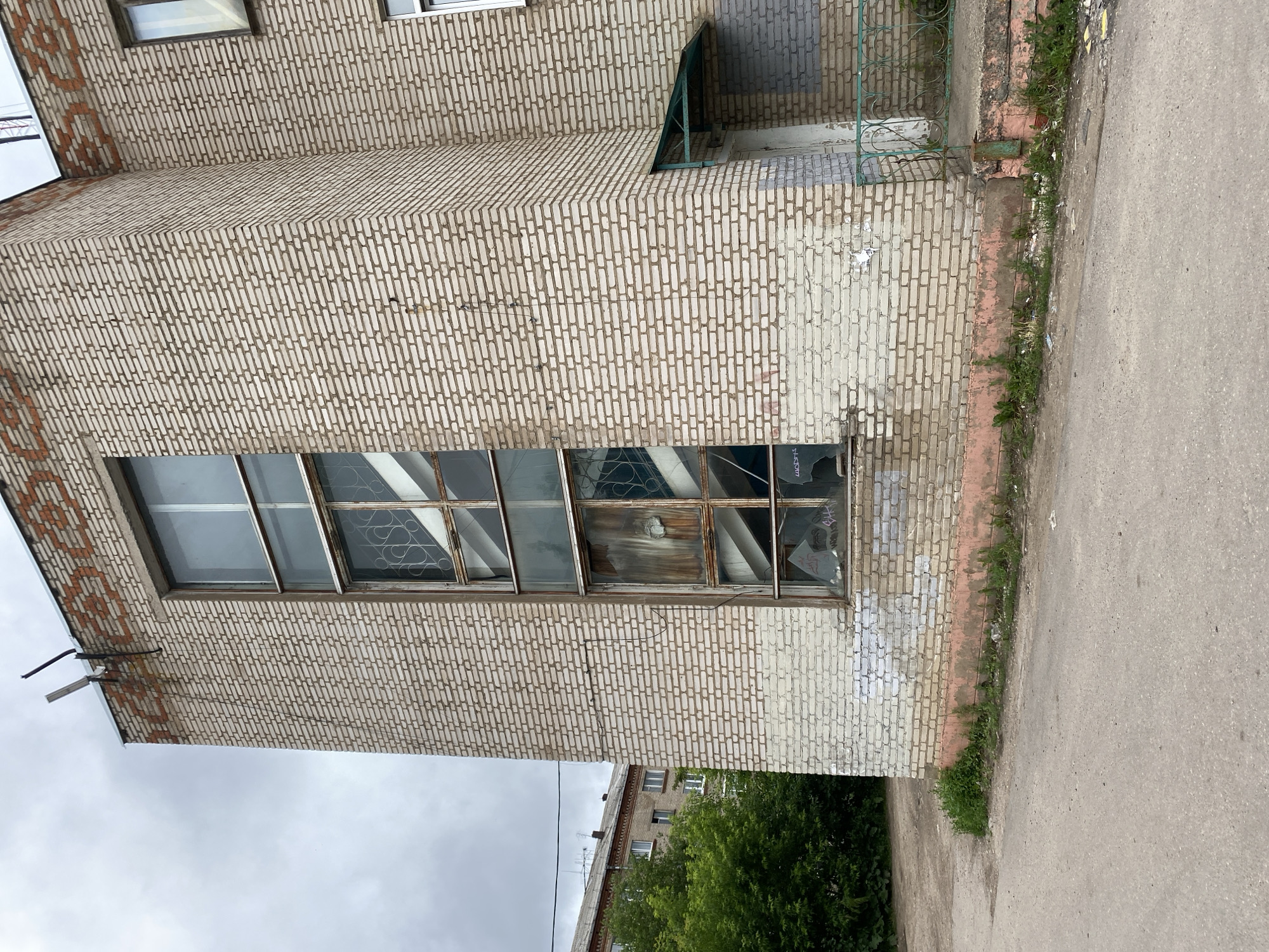 The image depicts the exterior of a multi-story brick building in Sergiyev Posad, Russia. The building's facade is constructed of light-colored bricks, with some areas showing signs of wear and graffiti. A tall, narrow window runs vertically along the side of the building, featuring a distressed frame and broken glass panes. Metal grates are visible behind the broken glass, suggesting a former security or decorative feature. A satellite dish is mounted on the exterior wall near the broken window. To the left of the building, lush green trees and another apartment building can be seen in the background. The foreground consists of an asphalt road and a small patch of grass with weeds growing along the building's foundation. The sky is overcast, indicating a cloudy day.