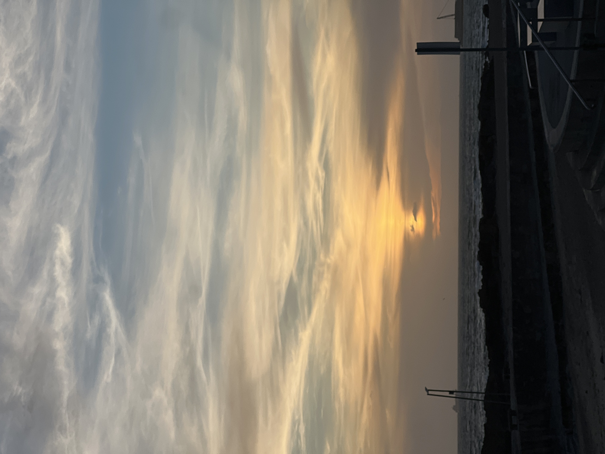 The image shows a sunset over the ocean in Porto, Portugal. The sky is filled with clouds, ranging from white and light blue on the left to golden and orange on the right. The sun is partially obscured by clouds, casting a warm glow. The ocean is visible as a dark strip. A stone wall with railings and steps is on the right side of the frame.