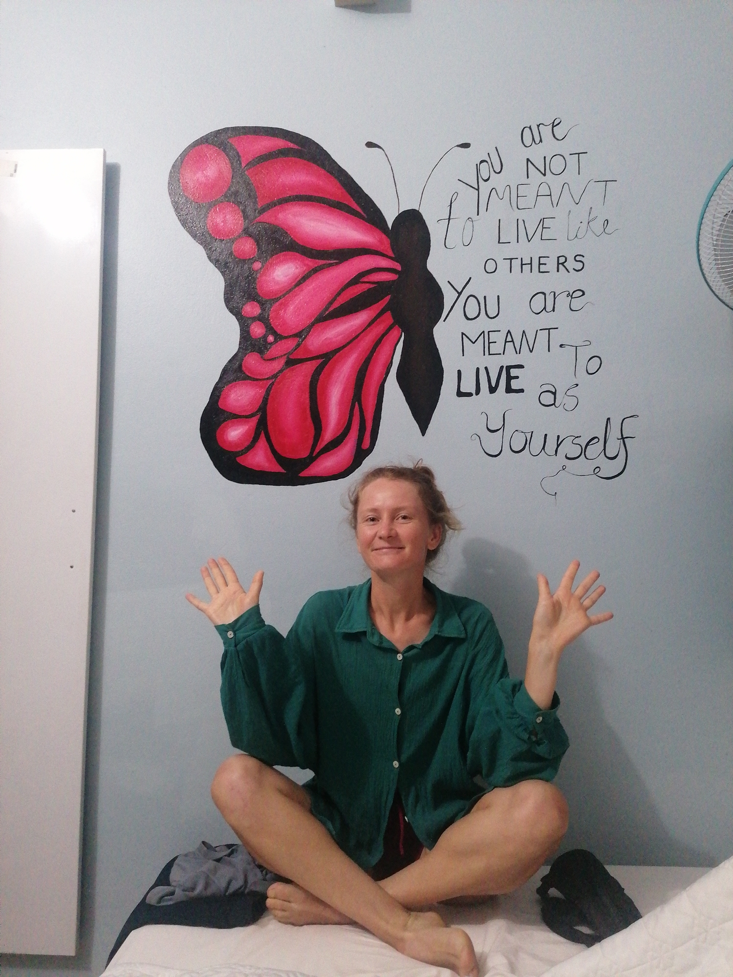 A fair-skinned woman with blonde hair is sitting cross-legged on a bed in a room, with her hands raised in a welcoming gesture. The wall behind her is painted light blue, and features a mural of a butterfly wing and text that reads, "You are not meant to live like others. You are meant to live as yourself." To the left of the woman is a white vertical panel, possibly a wardrobe or door. The bed is covered with a white sheet, and a pile of gray clothing and a pair of dark-colored shoes are visible on the bed next to her. The location is in Bai Chay Ward, Vietnam.