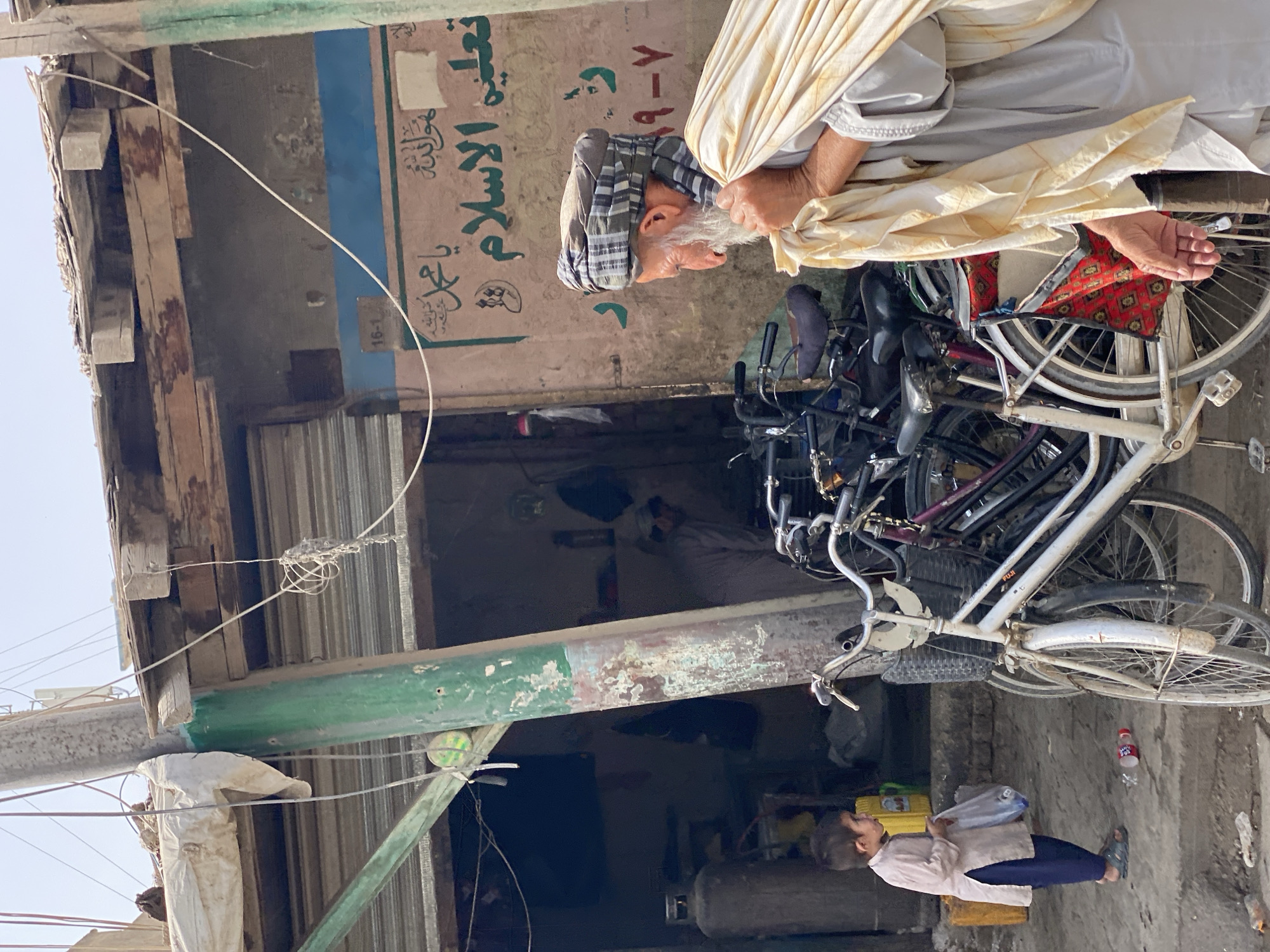 The image shows a street scene in Kandahar, Afghanistan. An elderly man, wearing a turban and traditional clothing, carries a bundle of fabric over his shoulder. He is standing in front of a building with Arabic script on the wall, which appears to be a shop or business. In the foreground, a young boy in a long shirt and pants stands holding a plastic bag. Several bicycles are parked on the street, suggesting a mode of transportation common in the area. The environment appears somewhat dusty and worn, with a concrete ground and a wooden structure overhead. The overall impression is of a bustling, everyday scene in a Middle Eastern city.
