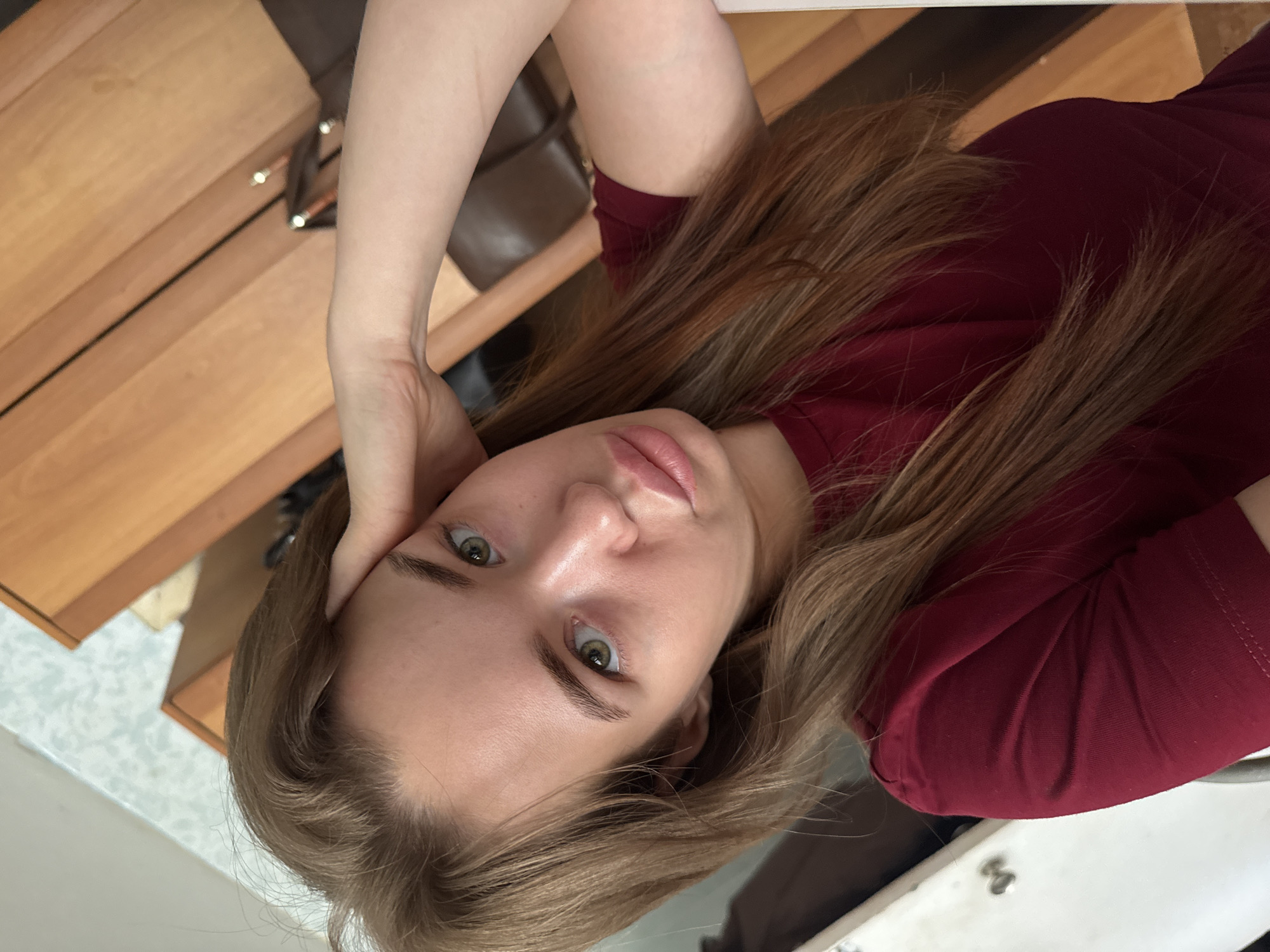 A young woman with long, light brown hair and green eyes is taking a selfie in an indoor setting. She is wearing a dark red t-shirt and has her left hand resting on her forehead. Her lips are slightly pursed, and she is looking directly at the camera. The background appears to be a room with wooden furniture and possibly a wall with patterned wallpaper. The lighting suggests it is daytime. There is no information to indicate the specific location within Chaikovsky, Russia.