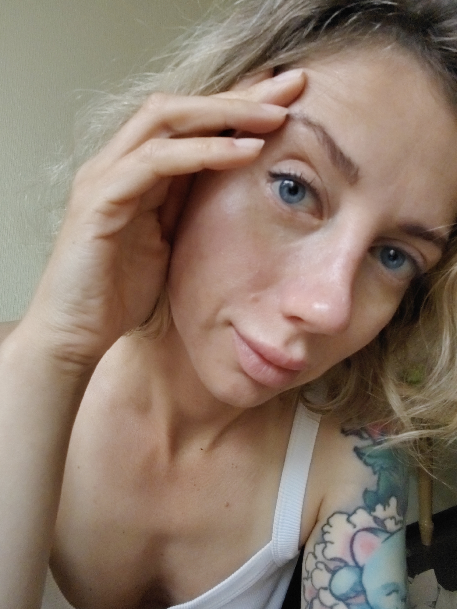 This is a close-up, selfie-style photograph of a woman with fair skin, blonde hair, and blue eyes. She is looking towards the camera, with her right hand touching her forehead and temple. Her expression is neutral.
She is wearing a white tank top. A colorful tattoo, featuring what appears to be an elephant and floral elements, is visible on her left shoulder and upper arm.
The background is softly lit and appears to be an indoor setting with a textured wall. The lighting suggests it could be daytime, with diffused natural light. There is no visible text in the image. The context provided places the location as Saint Petersburg, Russia.