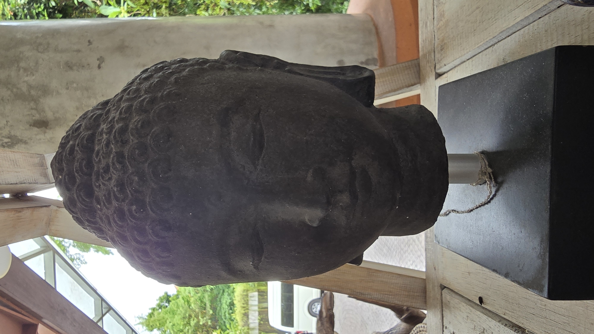 The image depicts a stone head of Buddha, resting on a black square base. A metal rod connects the head to the base, secured with a rope. The head is weathered and shows detailed curls on the head.
The scene appears to be indoors, with views of greenery, trees, and a partial view of a vehicle visible through windows in the background. The setting seems to be in Ubud District, Indonesia. The overall composition suggests a contemplative or display setting.