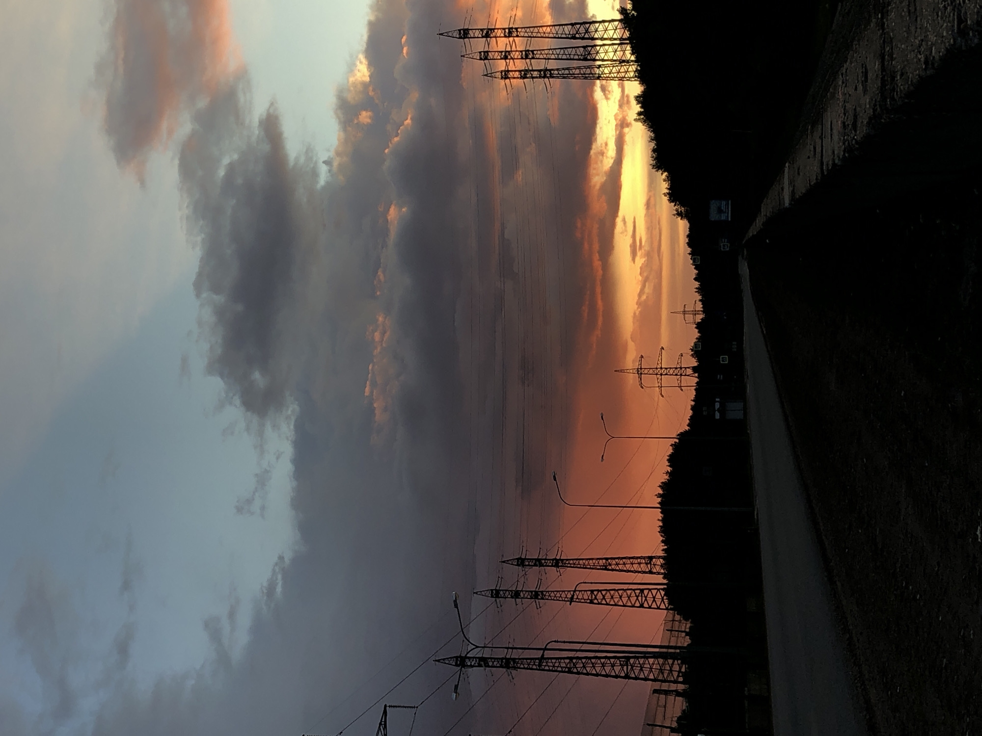 The image depicts a landscape at dusk in Noviy, Russia. The sky is filled with heavy, dark clouds on the left transitioning to shades of orange and yellow on the right, suggestive of a sunset. There are electrical power towers silhouetted against the colorful sky and along the side of the road. Streetlights are also visible among the power towers. A paved road extends into the darkness along the right side of the frame, bordered by dark foliage and trees. The overall scene is dimly lit due to the fading light and the shadows cast by the objects and foliage.