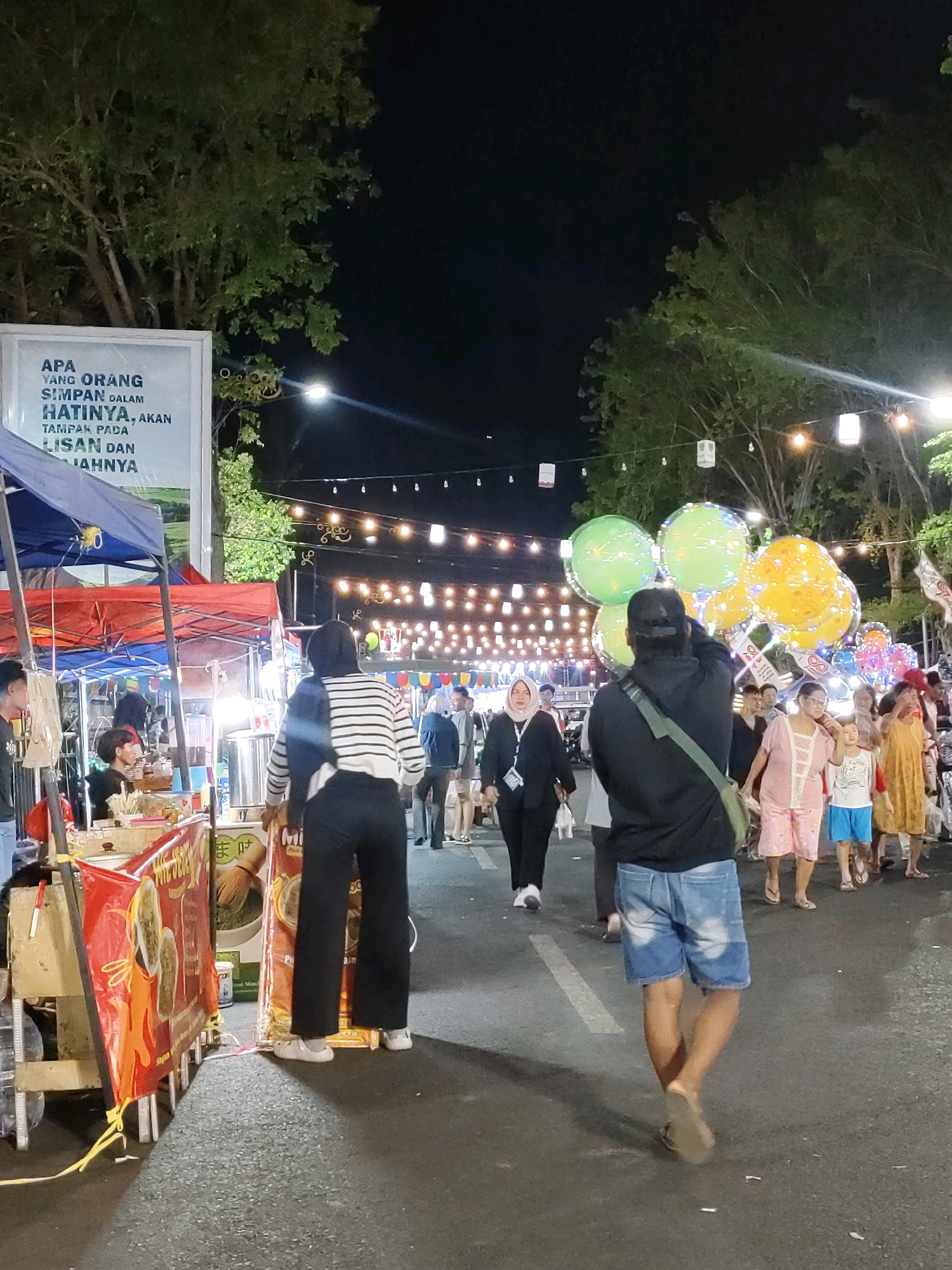 The image depicts a nighttime street scene in Probolinggo, Indonesia, likely a night market or a public gathering. Several stalls are set up along a paved street, with trees and string lights overhead. In the foreground, a person wearing a black hoodie and denim shorts walks away from the camera, carrying a green bag. In front of them, a group of people are holding a cluster of colorful balloons, some green and some yellow. To the left, a food stall is visible with red and yellow banners. Several other individuals are browsing the stalls or walking along the street. The atmosphere appears lively and social.