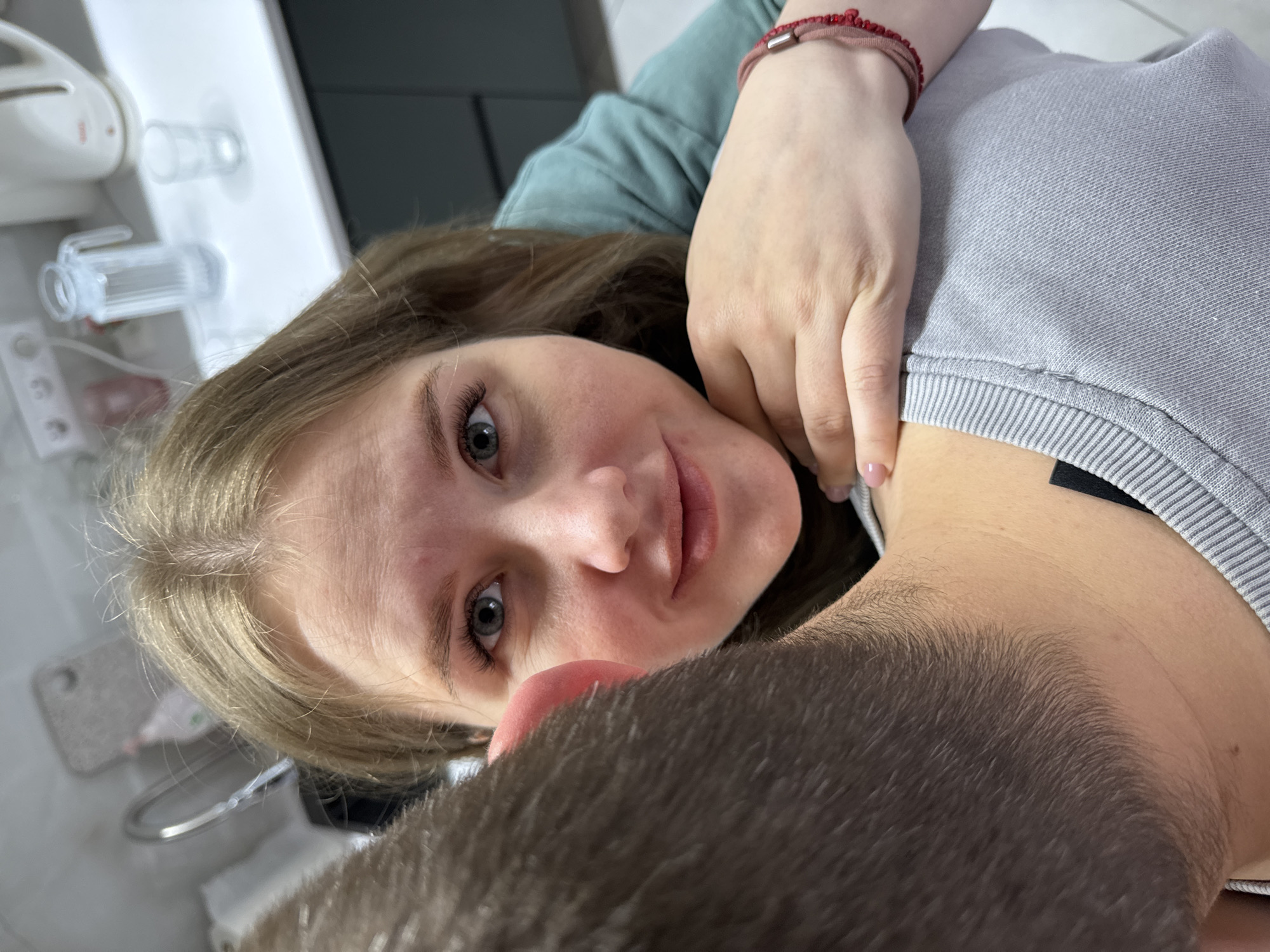 This close-up, angled portrait captures two individuals indoors in Gornyi (Dvurechenskoe S/P), Russia. The primary subject is a young woman with long, light brown hair and striking blue-grey eyes, looking directly towards the camera with a calm, slightly smiling expression. She is wearing a teal-colored long-sleeved top and a red beaded bracelet with a silver element on her left wrist. Her right hand is gently placed on the shoulder or chest of a man, whose head and short brown hair are visible in the lower right foreground, partially obscuring the view. He is dressed in a light grey sweater with ribbed cuffs, and a black strap (possibly from an undershirt or garment) is visible beneath his collar. The background features a white surface, likely a countertop or shelf, on which a white electric toothbrush and several clear plastic cups are placed, suggesting a bathroom or kitchen setting. The image evokes a sense of intimacy and affection between the two people. The lighting is soft and ambient, characteristic of an indoor environment. There is no visible text in the image.