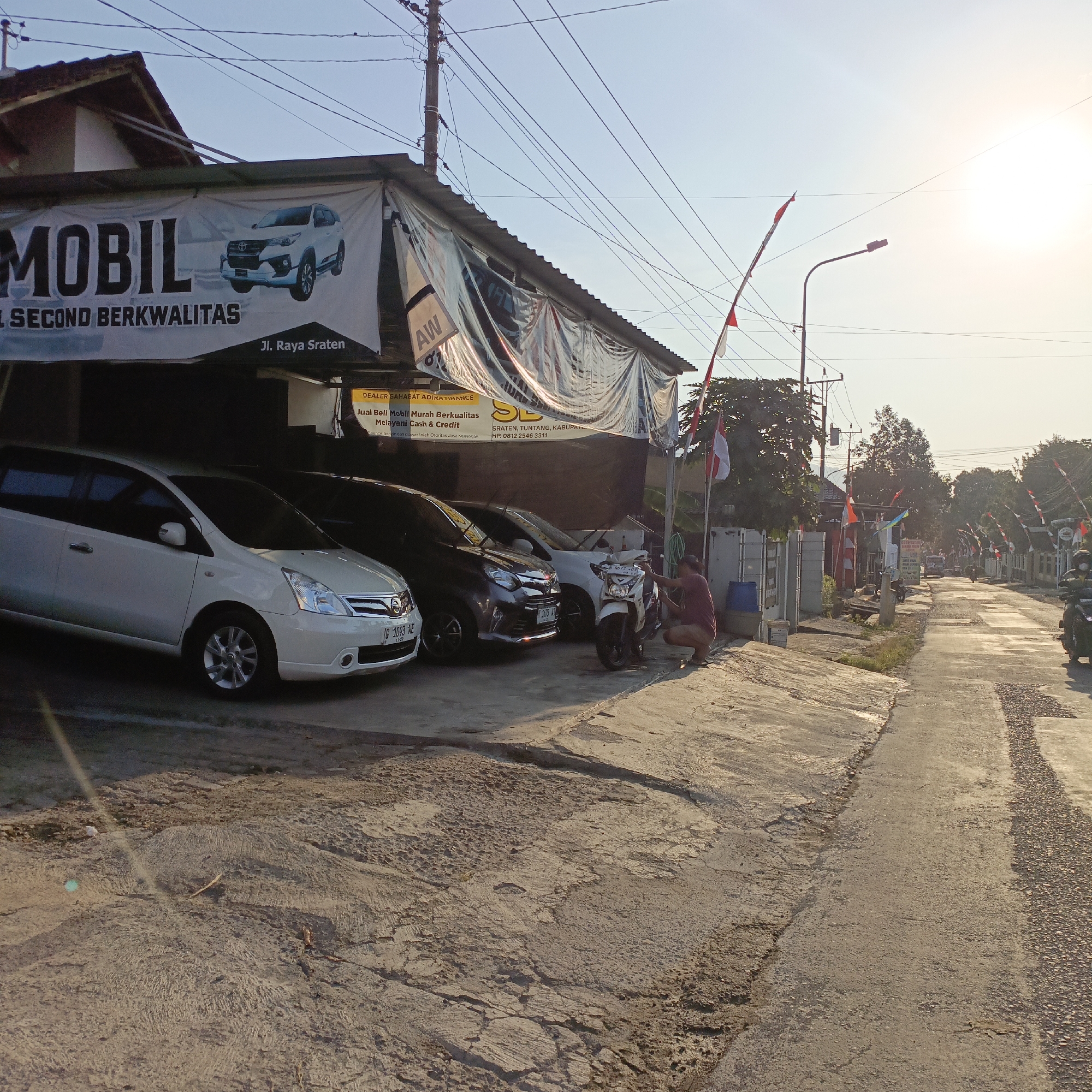 The image depicts a street scene in Sraten, Indonesia, under the bright glare of the sun. In the foreground, a white Nissan Grand Livina is parked alongside other vehicles. A man is crouched beside a white motorcycle, seemingly engaged in some activity. Behind the cars, a banner advertises "MOBIL JUAL BELI SECOND BERKUALITAS" (Used Cars for Sale, Good Quality) and "Jl. Raya Sragen" (Sragen Raya Street), indicating the location of a used car dealership. Red and white Indonesian flags are visible on poles along the street, suggesting a festive or commemorative occasion. The street itself appears to be paved with concrete and asphalt, showing signs of wear and tear. Power lines crisscross the sky, and street lights are present. The overall impression is a glimpse into a daily scene in a Southeast Asian town, with commerce and community elements intertwined.