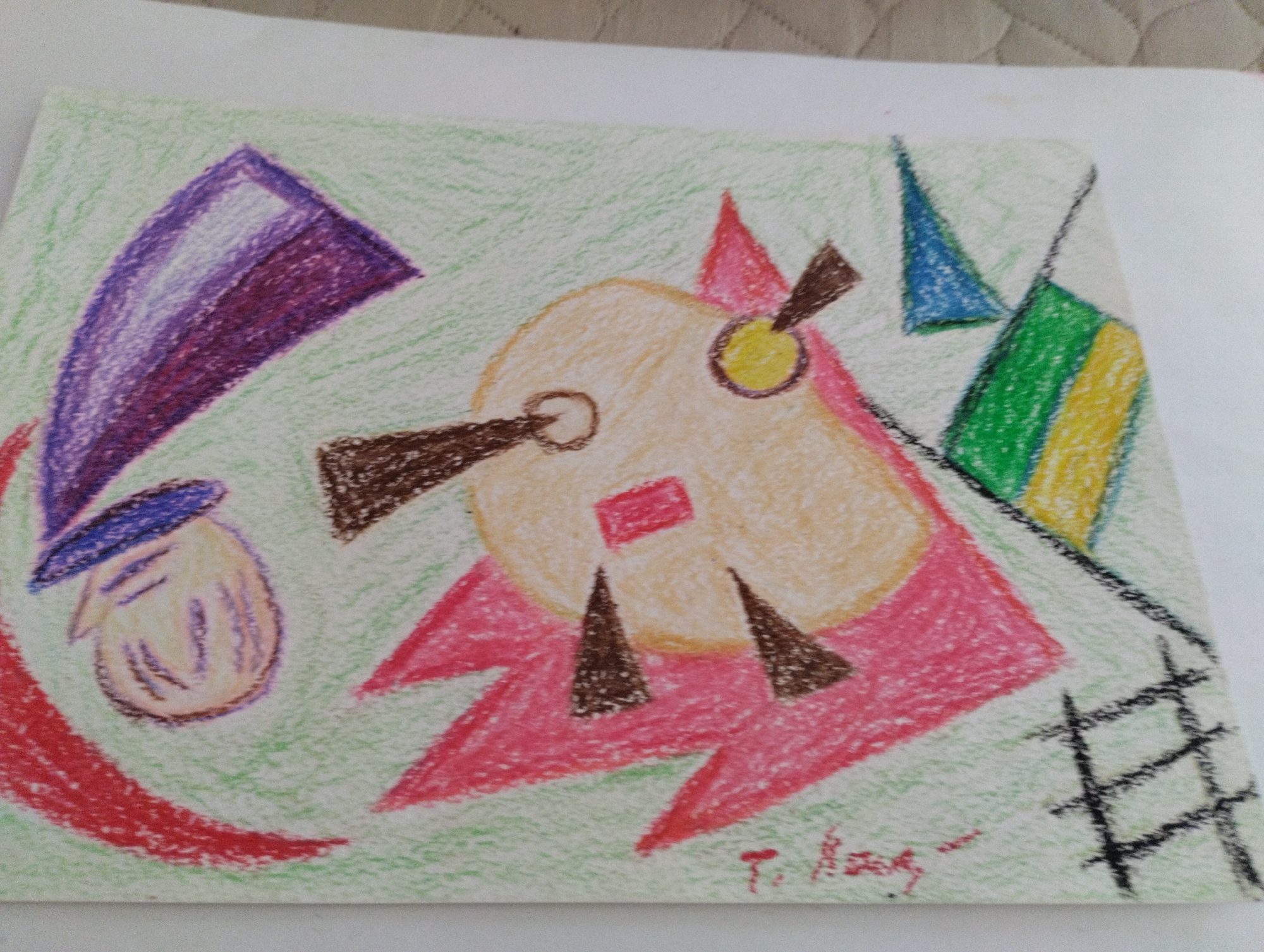 This is a photograph of an abstract drawing created with colored pencils on white paper. The drawing features a central circular shape with a tan-colored fill, outlined in pink. Two brown triangular shapes point upwards from below this circle, and a smaller red square is centered on its upper portion. To the left, a stylized head and hat are depicted, with a purple and white hat and a tan face. A red curved line forms a base on the left side. To the right of the central circle, there are abstract shapes including a blue triangle, and a green and yellow rectangular shape, all rendered in colored pencil. A black grid-like structure is present in the bottom right corner. The drawing is signed in red cursive at the bottom right. The paper is placed on a textured surface, likely a tablecloth or a woven mat. The context of Mezitli, Turkey is not depicted or suggested by the image itself.