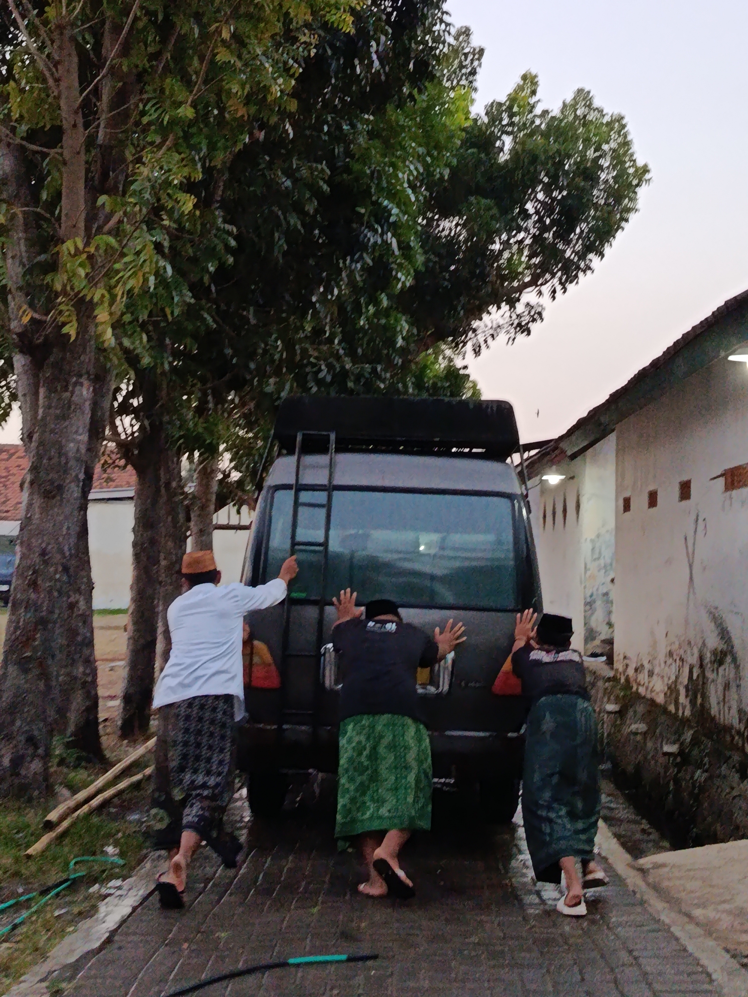 In the city of Ramban Wetan, Indonesia, three individuals are pushing a dark-colored van backward on a paved surface. The van has a ladder attached to its rear roof rack. The individuals are dressed in traditional Islamic attire, including peci (caps) and sarongs. One person on the left wears a white long-sleeved shirt and a patterned sarong, while the other two on the right wear dark t-shirts and solid-colored sarongs. The scene is situated outdoors, with large trees on the left side of the image and a building with white walls on the right. The sky appears overcast or at dusk. A green hose is visible on the ground to the left of the van.