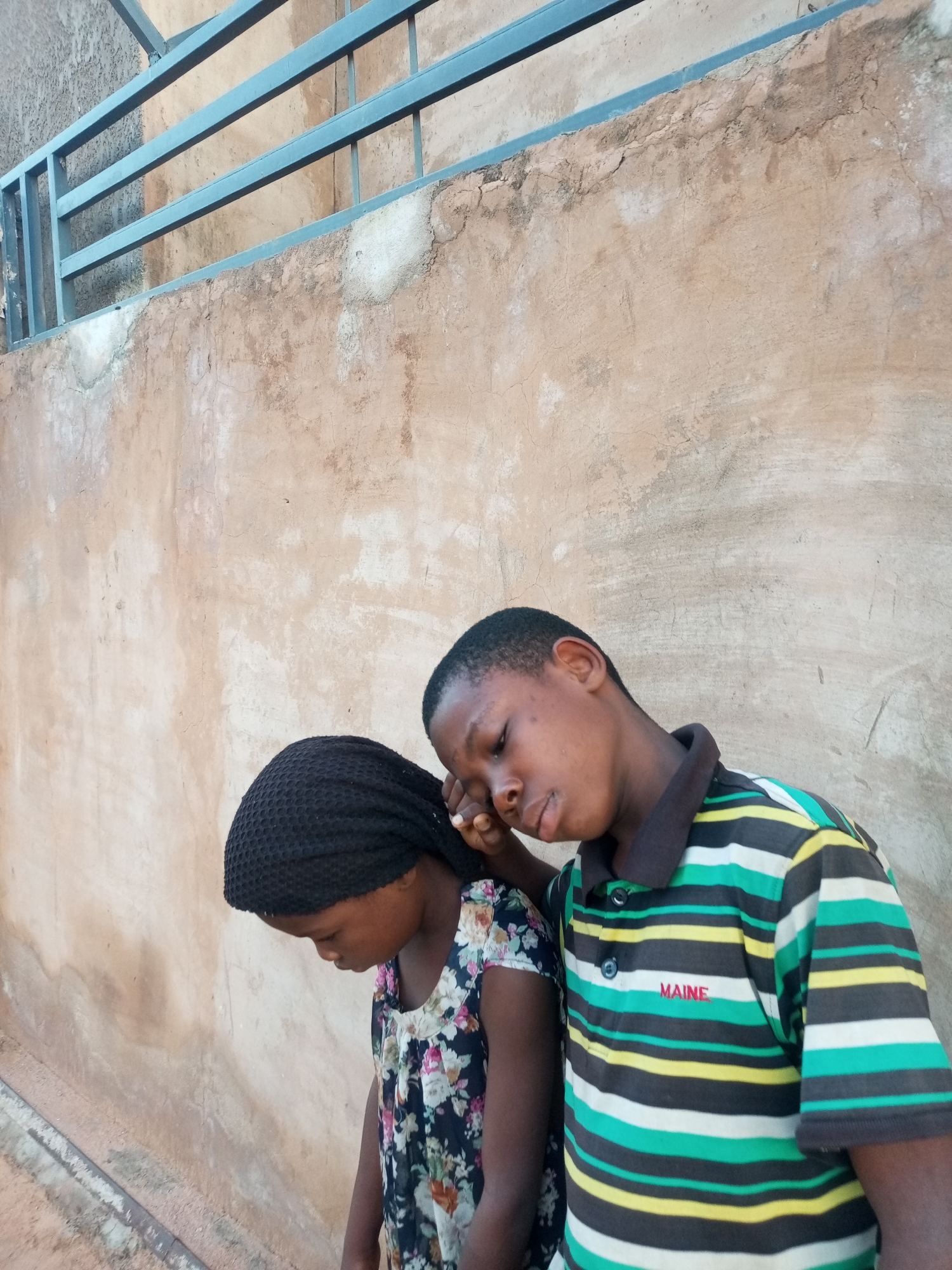 The image captures two young Black individuals, a boy and a girl, standing beside a textured, light brown wall that appears to be part of an outdoor setting in Tempure, Nigeria. A metal railing, painted a dull blue, runs horizontally across the top of the frame, above the wall.
The girl, on the left, is wearing a black headscarf and a floral-patterned dress with a dark base and colorful flowers. Her head is bowed, and her gaze is directed downwards, obscuring her facial features.
The boy, positioned to the right and slightly behind the girl, is wearing a horizontally striped polo shirt in shades of green, white, yellow, and black. A small red embroidered logo that appears to read "MAINE" is visible on the left chest of his shirt. His eyes are closed, and his head is tilted, with his right hand gently resting on the girl's head or shoulder, suggesting a supportive or comforting gesture.
The lighting in the image is somewhat diffused, indicating it might be overcast or taken during a time of day without harsh sunlight. The wall exhibits signs of weathering and age, with some patches of lighter material and fine cracks visible on its surface. The overall scene is still, with the focus on the two children and their interaction against the backdrop of the weathered wall and railing.