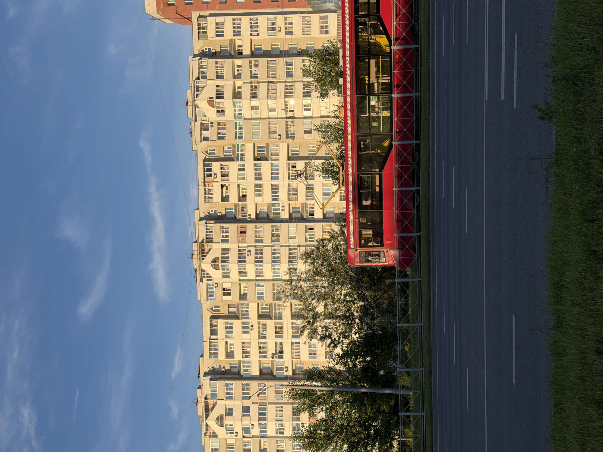 The image depicts a street scene with a red tram on the right side of the frame, moving along a paved road marked with white lines. A tall, light-colored apartment building dominates the left side of the image, featuring numerous windows and balconies. Trees with green foliage are visible between the tram and the apartment building, partially obscuring the lower floors of the building. The sky is clear and blue with a few wispy clouds, suggesting it is daytime. The lighting indicates it might be late afternoon or early evening, with the sun casting a warm glow on the building. The scene appears to be in an urban environment, likely a residential area. No people are clearly visible in the image, but the presence of a tram suggests a public transportation route. There is no visible text in the image.