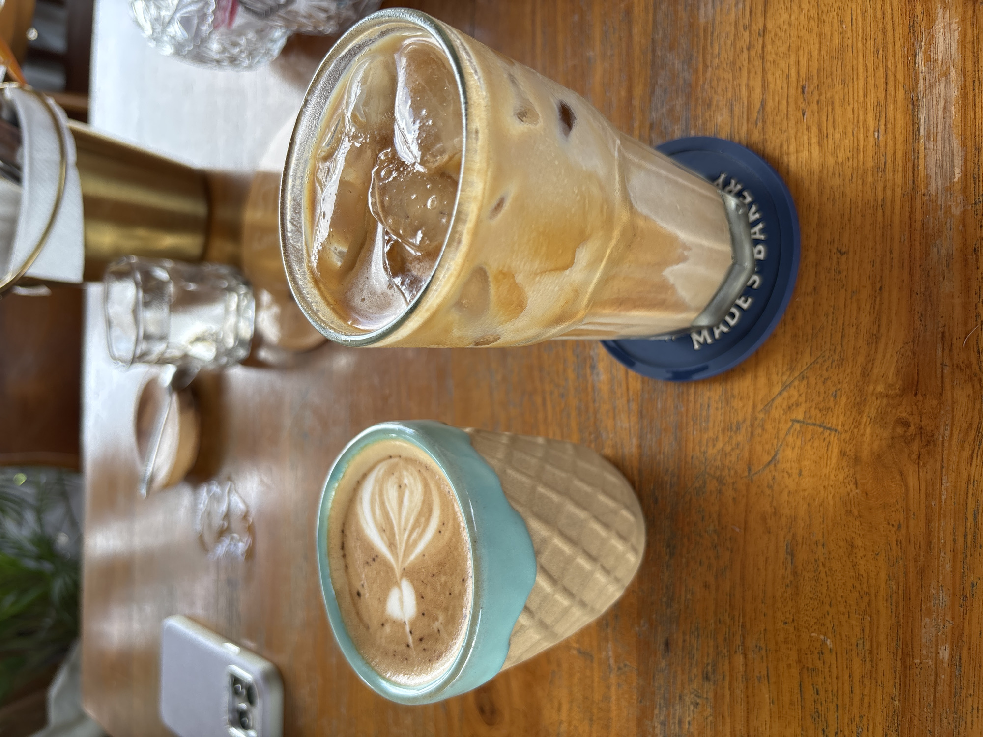 An overhead shot captures two inviting beverages presented on a wooden table, likely within a cafe setting in Kuta Selatan, Indonesia. In the foreground, a unique ceramic cup, styled like an ice cream cone with a textured waffle pattern and a distinctive light blue rim, holds a hot latte. The latte features delicate white latte art, resembling a heart or a tulip.
Adjacent to it, a tall, clear glass contains an iced coffee, brimming with ice cubes and a creamy, light brown coffee mixture. This glass rests on a round, dark blue coaster, which clearly displays white embossed text reading "MADES GALLERY." Partially visible in the bottom left corner of the frame is a smartphone in a light-colored, possibly grey or lavender, case, lying on the table.
The wooden table exhibits a natural grain and some signs of wear. The background is softly blurred, revealing hints of other items such as a small clear glass, a metallic, possibly gold-toned, cylindrical object, and some green foliage, suggesting indoor plant decor. The lighting is bright and even, highlighting the fresh appearance of the drinks. No people are visible in the image.