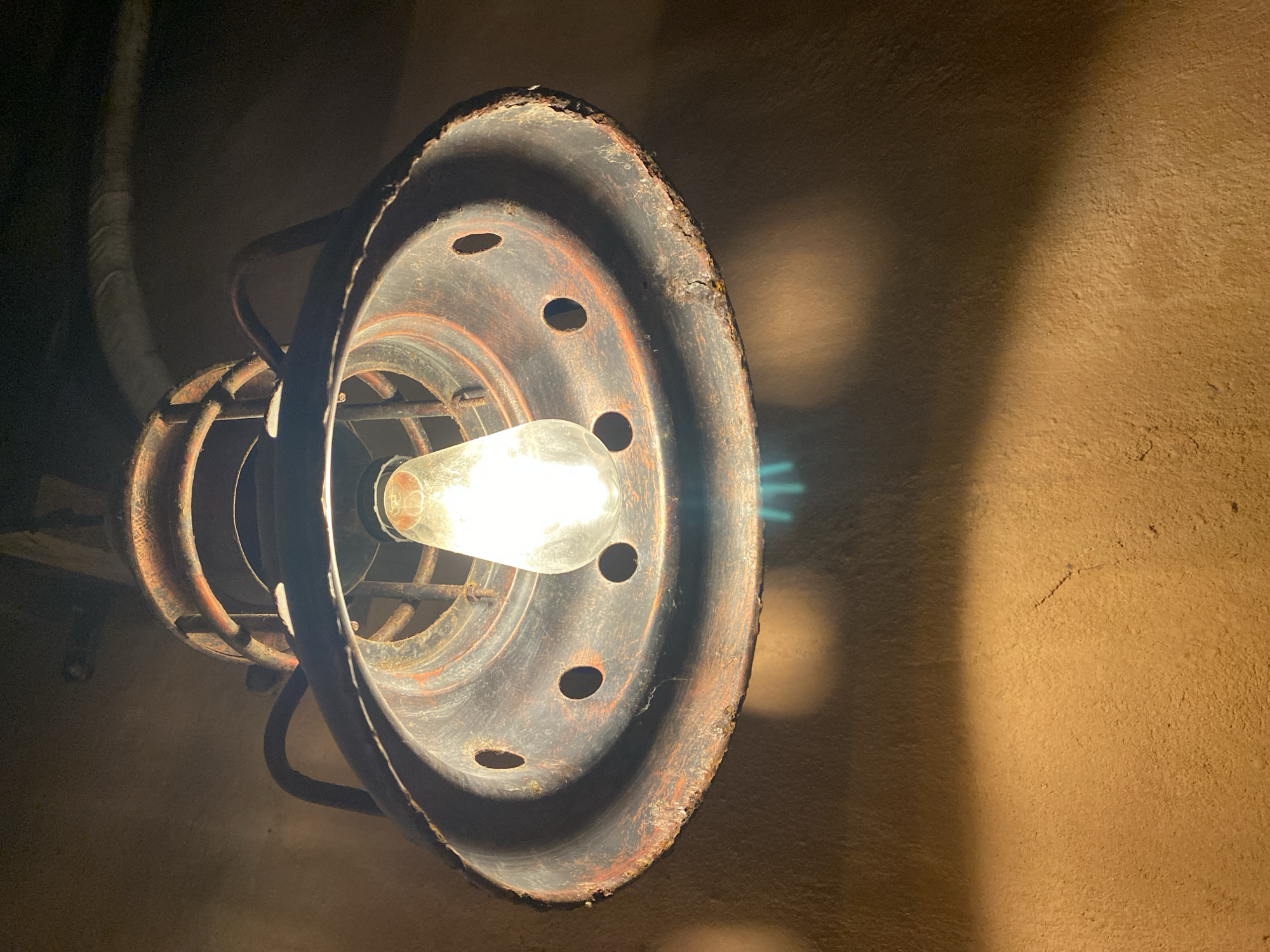 The image shows an industrial-style pendant light fixture with a visible Edison bulb, casting light downwards onto a textured, earthy-colored wall. The fixture appears to be made of dark metal, possibly rusted, with a cage-like enclosure around the bulb and a wide, flared shade with several circular openings. The wall has a rough, uneven surface and is illuminated by the light, creating a warm glow and shadows. The scene is dimly lit overall, with the primary light source being the pendant lamp. There is no indication of people or specific actions. The location in Đà Nẵng, Vietnam, cannot be determined from the visual information provided in the image.
