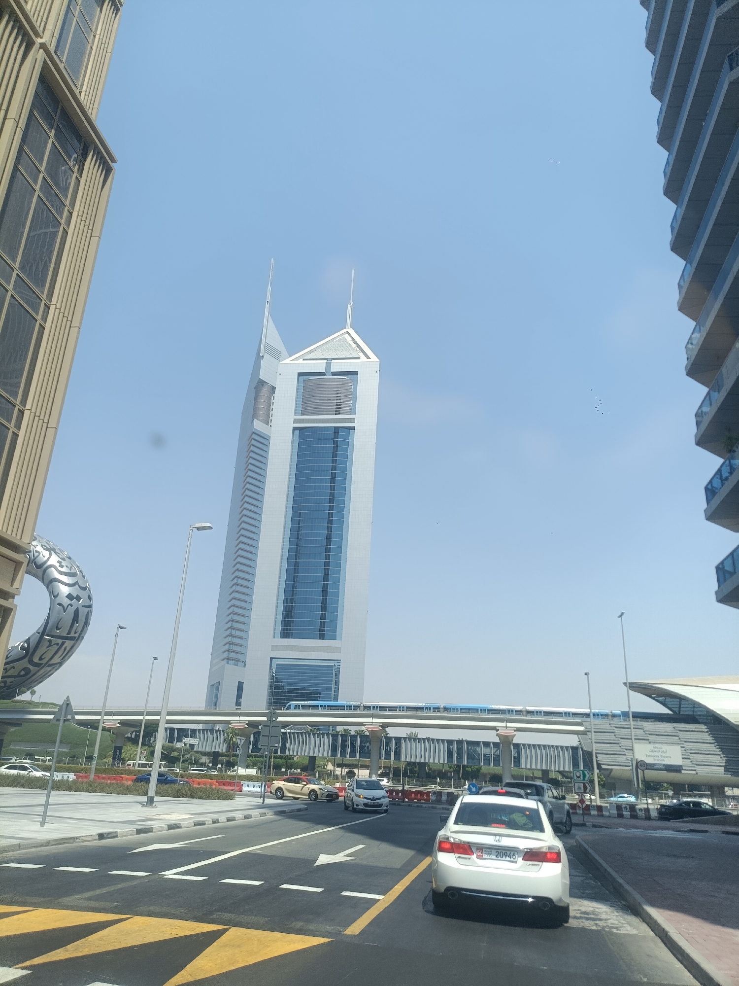 The image captures a street view in Dubai, United Arab Emirates, showcasing modern architecture and urban infrastructure under a clear blue sky. In the foreground, a white sedan is prominently visible, facing away from the camera, with its license plate reading "50 20946". The car is positioned on a multi-lane road marked with white directional arrows and yellow diagonal stripes. Other vehicles, including a silver SUV and a beige sedan, are also present on the road, indicating moderate traffic flow.
To the left of the road, a large, abstract metallic sculpture with Arabic calligraphy is partially visible, hinting at public art installations. Behind it, a section of a beige-colored building with a grid-like facade stands tall. Dominating the center of the image is a striking, slender skyscraper with a distinctive angled top, its facade a combination of white panels and blue glass. This skyscraper is identified as Emirates Towers.
In the middle ground, a raised metro track runs horizontally across the frame, with a blue metro train visible on it. The metro station, with its sleek, modern design, is situated on the right side of the image, featuring a covered platform and architectural elements that complement the surrounding skyscrapers. The environment appears clean and well-maintained, characteristic of Dubai's urban landscape. Streetlights are strategically placed along the road and sidewalks. The overall scene depicts a bustling urban environment with a focus on contemporary design and efficient public transportation.