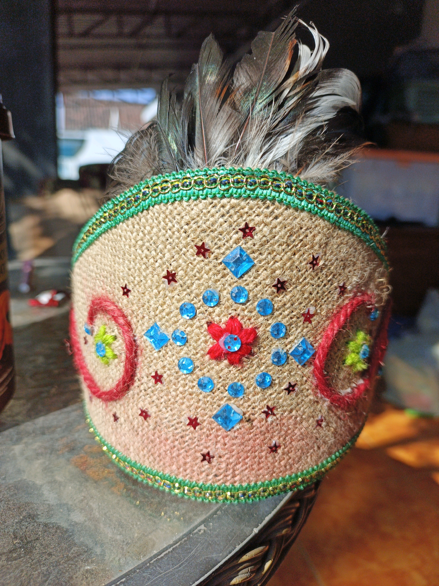 A close-up, eye-level shot shows a decorated headband with feathers. The headband is made of burlap-like material and adorned with red stars, blue square and circular sequins, and two embroidered circular designs. A green and gold trim runs along the top and bottom edges of the headband, and several grey and brown feathers are stuck into the top. The headband is resting on a weathered, grey surface that appears to be a table or ledge. In the blurred background, there is a glimpse of an outdoor setting with structures and sunlight. This image was taken in Ketegan, Indonesia.