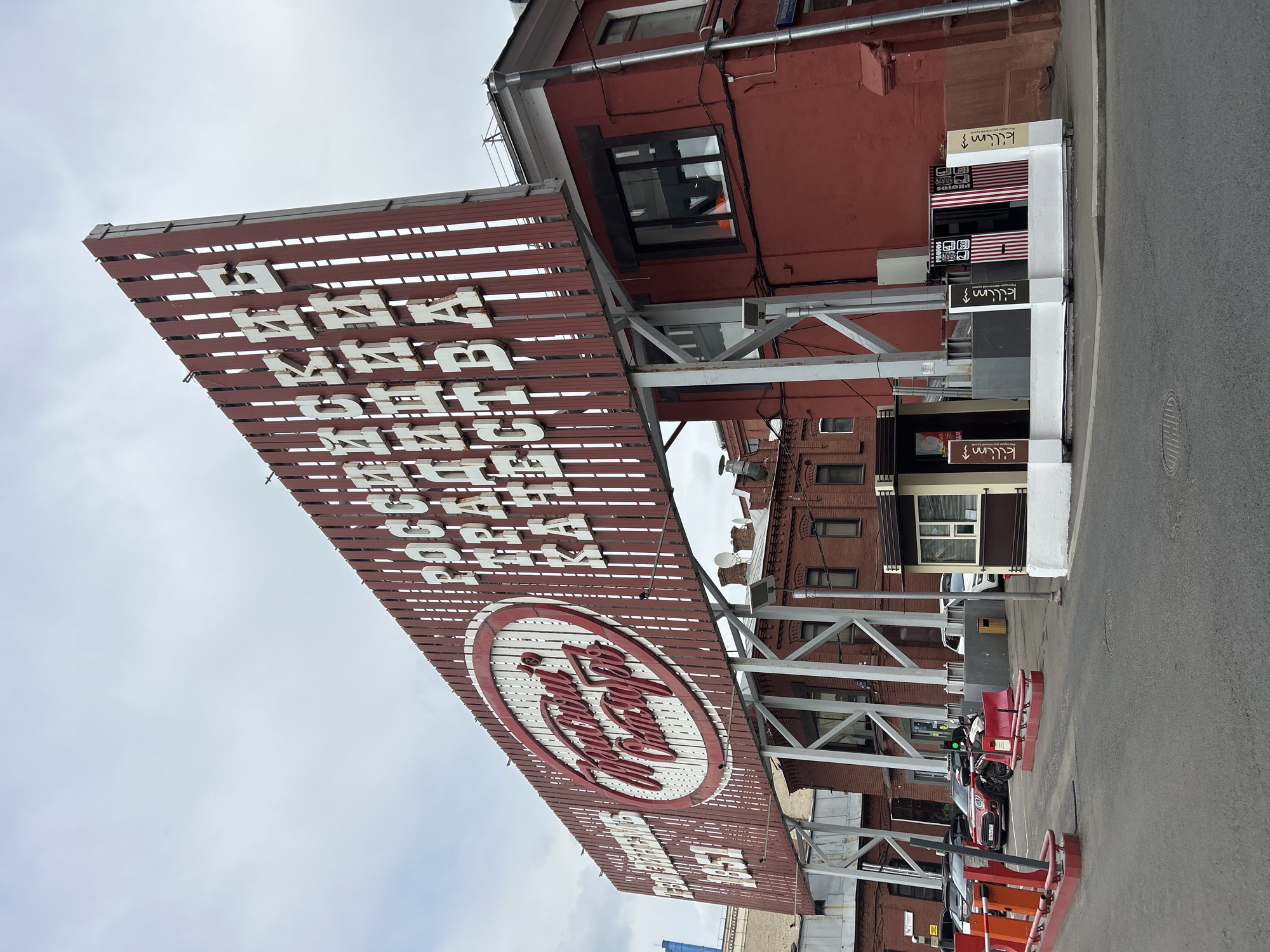 The image shows a large, weathered sign with white lettering on a brown wooden background, mounted on a steel frame structure. The sign includes a circular emblem with cursive writing inside. Above and below the emblem, additional lines of blocky lettering are visible. The sign is attached to the side of a red brick building with windows. Below the sign, a sidewalk, a street, parked cars, and a white-painted structure with signage are visible. The overall scene appears to be an urban environment. One of the signs reads "Kilim".