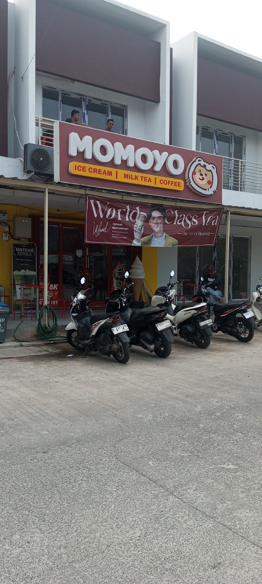The image shows the exterior of a Momoyo store in Setu, Indonesia. The store has a prominent red sign with the Momoyo logo and text indicating "ICE CREAM | MILK TEA | COFFEE". Below the sign, a large banner displays "World Class Tea" featuring a picture of Iqbal Ramadhan, an actor and musician, described as a "Momoyo Brand Ambassador".
In front of the store, several motorcycles are parked on the pavement. To the left, a green hose is coiled on the ground next to a trash can. An air conditioning unit is mounted on the building above the entrance. Two men are visible on the balcony of the building. One man is wearing a black t-shirt and the other a grey t-shirt and glasses. Another man, wearing a black jacket and a cap, is sitting on a motorcycle in the background, looking at his phone. The overall environment appears to be a commercial area with a sidewalk and street in front of the shops. The building is modern with white and brown elements.