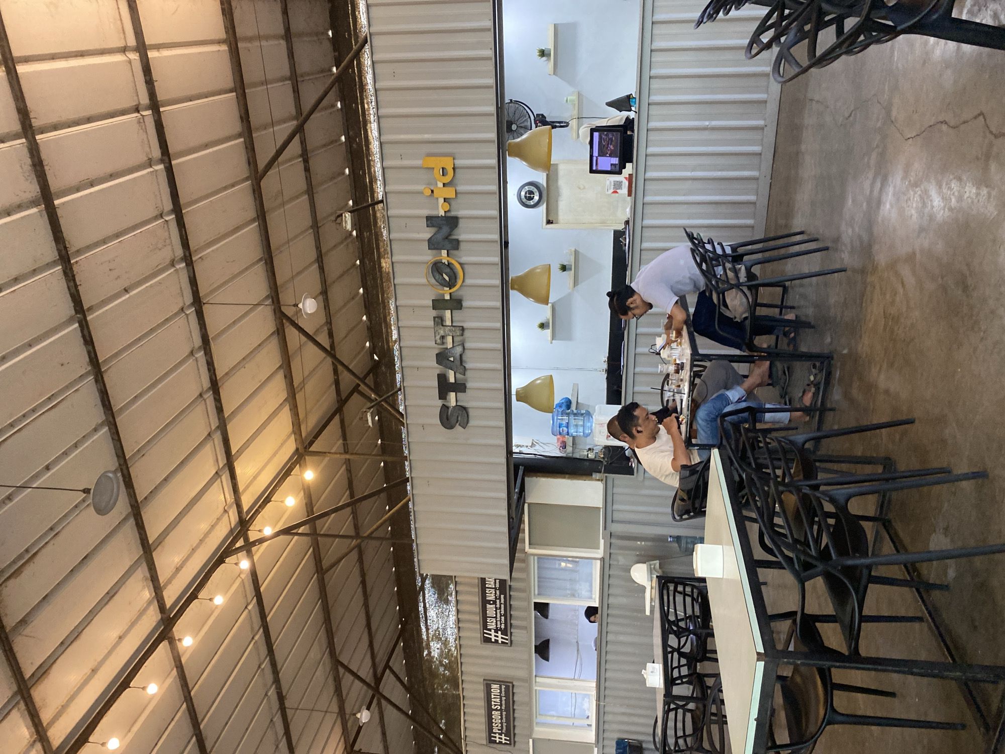 This image depicts the interior of a restaurant named "Pi-NOT-als," located in Lhokseumawe, Indonesia, as indicated by the sign stating "#PISGOR STATION" and "#NASI UDUK." Two men are seated at a table, interacting with food or objects on the table, possibly eating or paying. The restaurant's interior features a high, industrial-style ceiling with exposed beams and lighting. Several empty black metal chairs are arranged around tables, suggesting a waiting or unoccupied dining area. The walls appear to be constructed from corrugated metal panels.