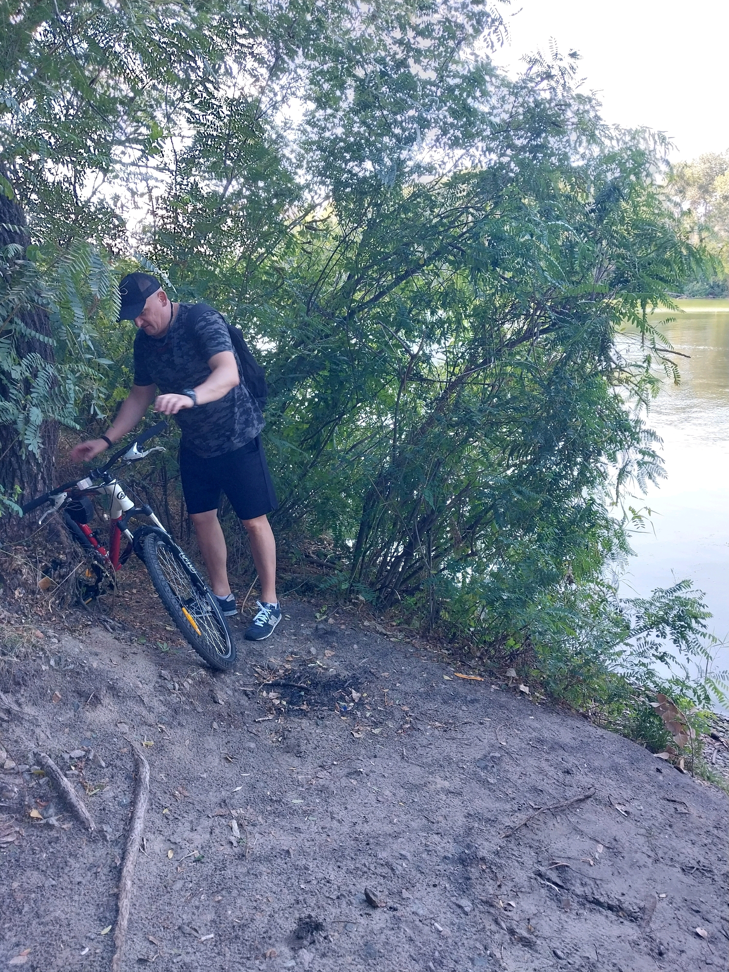 A man in a black t-shirt, black shorts, and a black baseball cap stands next to a bicycle on a dirt path near a body of water, likely the Dnipro River in Dnipro, Ukraine. The man is leaning down, appearing to adjust something on the bicycle, which is leaning against a tree. The bicycle has a red frame and black tires. Dense green foliage is visible behind the man and the bicycle, with the water of the river to the right. The ground is covered in dirt, leaves, and some roots. The lighting suggests it is daytime, possibly late afternoon given the angle of the light filtering through the trees. The overall scene depicts a moment of pause during an outdoor activity, possibly cycling or hiking, in a natural setting within the city.
