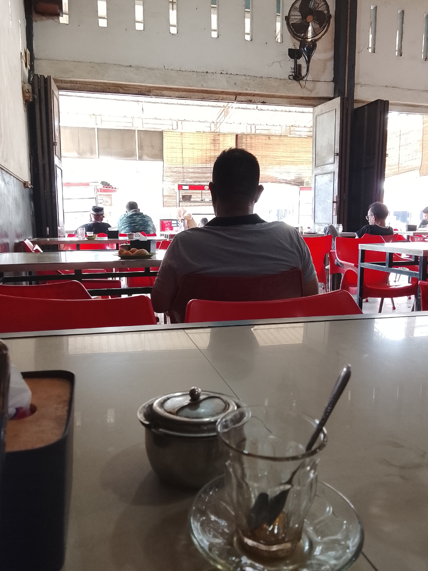 The image captures an interior view of a restaurant or cafe in Lhokseumawe, Indonesia. The foreground features a table with a sugar bowl, a glass with a spoon, and a tissue box. Several people are seated at tables in the background, most with their backs to the camera. Red plastic chairs are visible around the tables. The restaurant appears to have large open doorways or windows, providing a view of the outside and allowing natural light to enter. A wall-mounted fan is visible near the ceiling.
