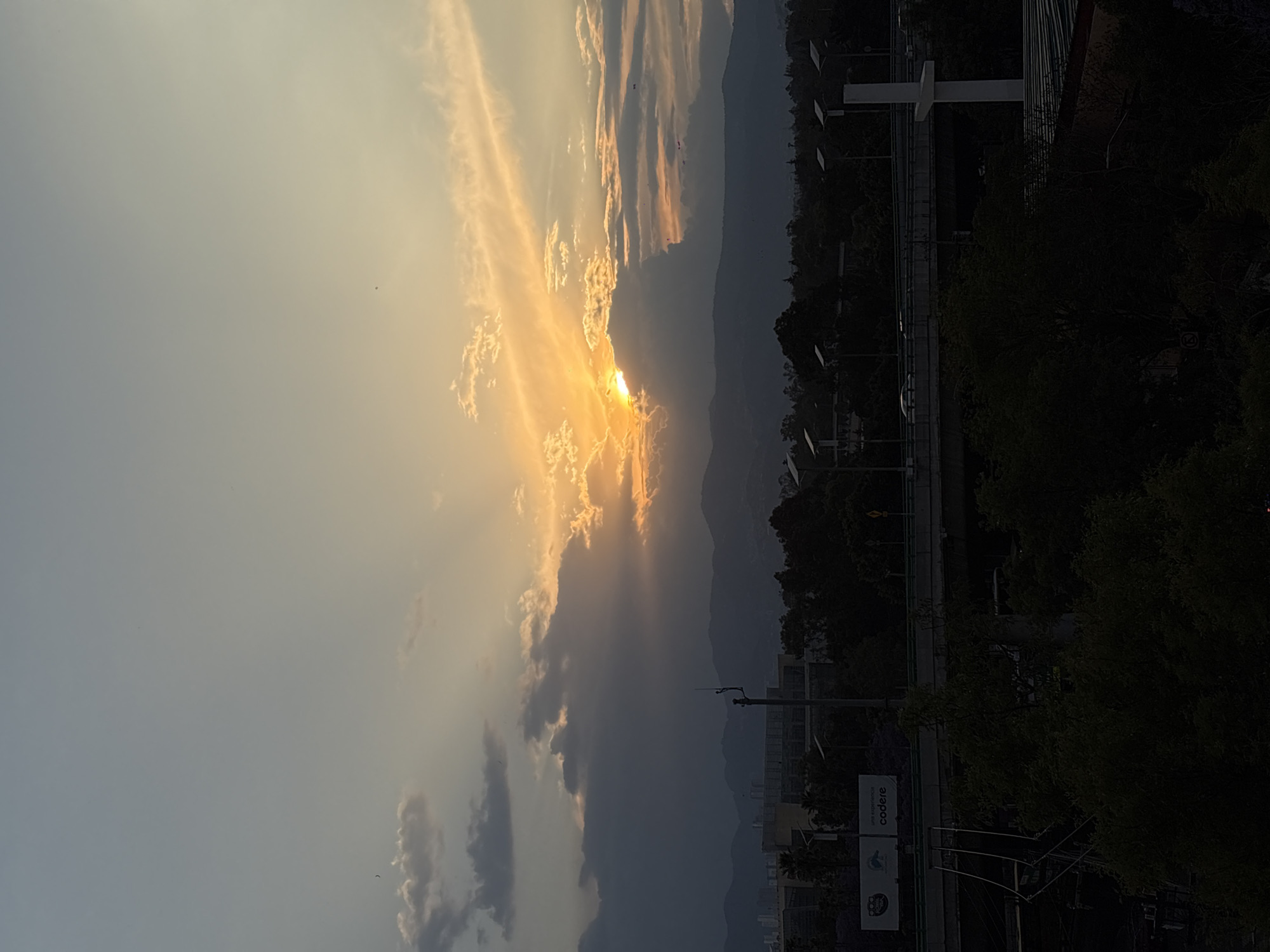 The image shows a sunset in Mexico City, Mexico. The sun is partially obscured by clouds, with rays of light streaming through the gaps. In the background, there are mountains. A highway with lamp posts runs through the middle ground. In the foreground, trees and buildings are visible. A billboard advertising "codere" can be seen near the bottom of the image.