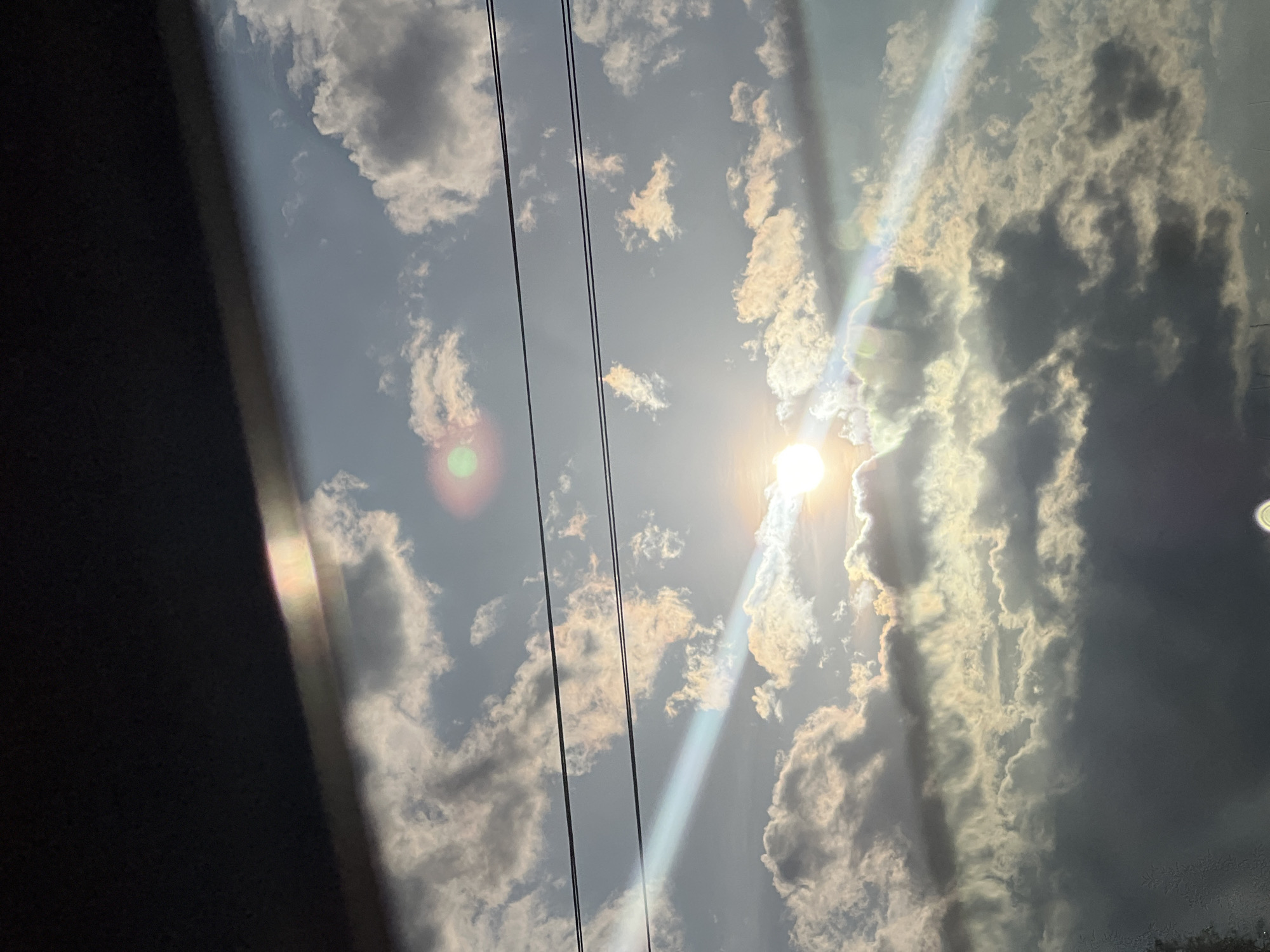 The image captures a bright, partly cloudy daytime sky. The sun is prominently visible on the right side of the frame, casting intense light and creating significant lens flares and sunbeams that stretch across the sky. Clouds are scattered throughout the frame; those directly illuminated by the sun appear bright white and golden, while others are darker gray, indicating varying density or being in shadow.
Three dark, thin lines, identifiable as power lines, traverse diagonally from the upper left to the lower right of the image. On the far left, a substantial dark area with a slightly lighter, irregular edge suggests a window frame or a structural element of a vehicle or building, from which the photograph was taken, partially obscuring the view.
The scene depicts a sunny but partly cloudy day, likely during late morning or afternoon due to the strong, direct sunlight. The visible power lines suggest an outdoor environment with infrastructure. The image provides a serene view of the sky from Pesnopoy, Bulgaria, without any visible text or human subjects.