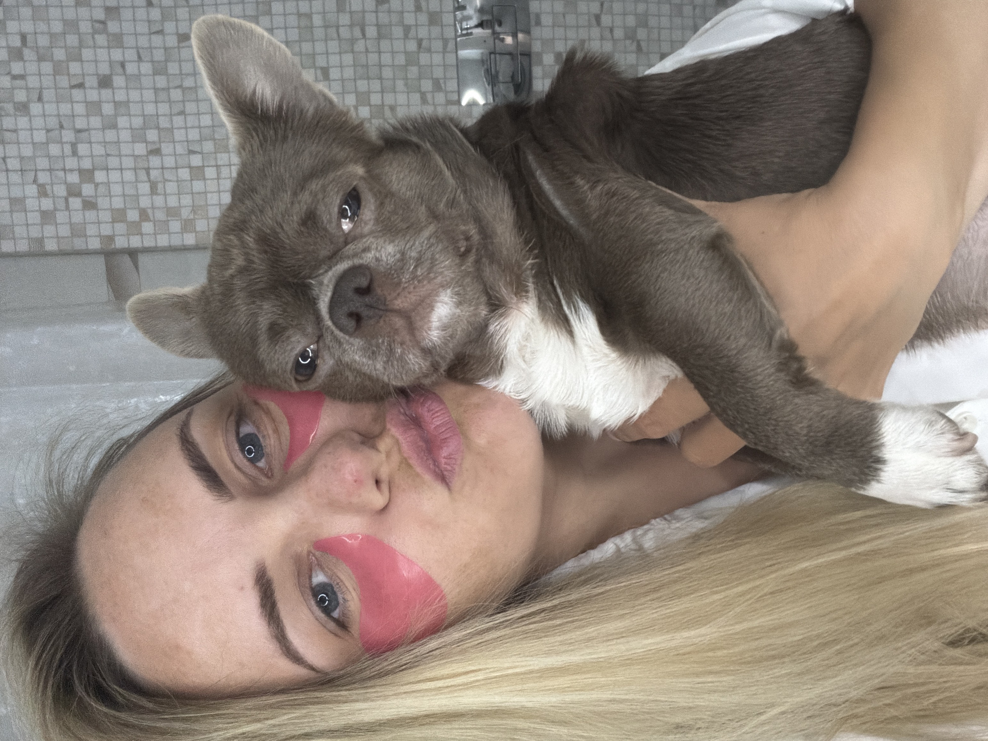 This close-up indoor shot, likely taken in Votkinsk, Russia, features a woman and a small dog in a relaxed, affectionate pose. The woman, with blonde hair and fair skin, is lying down and has bright pink under-eye patches applied to her face, suggesting a skincare routine or moment of self-care. Her lips are slightly parted in a soft pout, and her eyes, looking directly at the viewer, convey a calm and gentle expression. She is cradling a small, grey-brown dog with distinct white markings on its chest and paws, possibly a French Bulldog or similar breed. The dog is resting its head close to the woman's face, also looking towards the camera with large, expressive eyes.
The setting appears to be a bathroom, indicated by a tiled wall with small, light-colored square tiles and a partially visible silver faucet in the background. The lighting is soft and even, typical of an indoor environment. The image captures a moment of intimate companionship and relaxation between the woman and her pet. No text is visible in the media file.