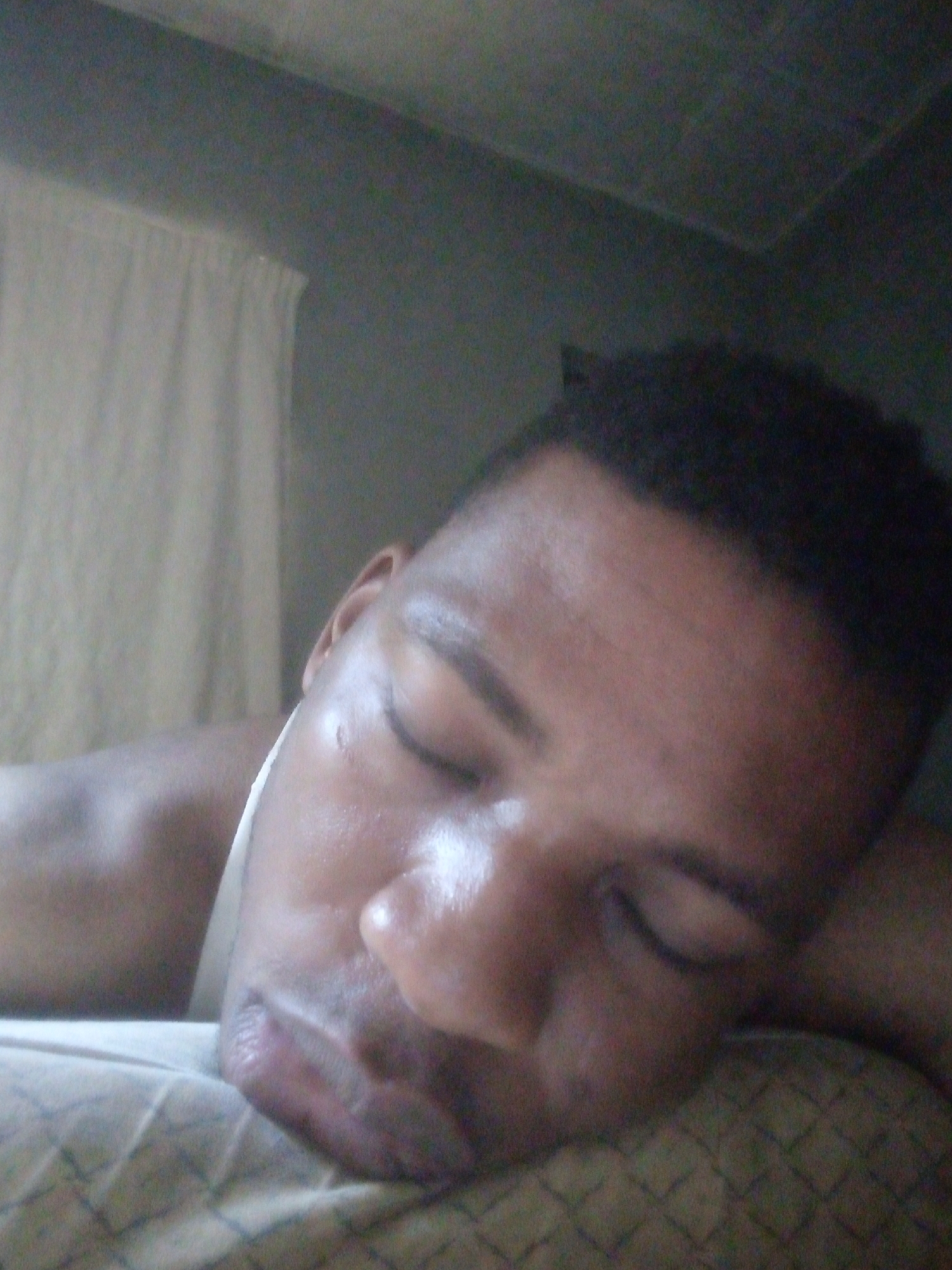 A close-up, medium-shot photograph shows a dark-skinned man lying on a pillow with his eyes closed. The man has short, dark hair. The pillow has a textured pattern. Behind the man are a curtain and a wall. The location is unknown.
