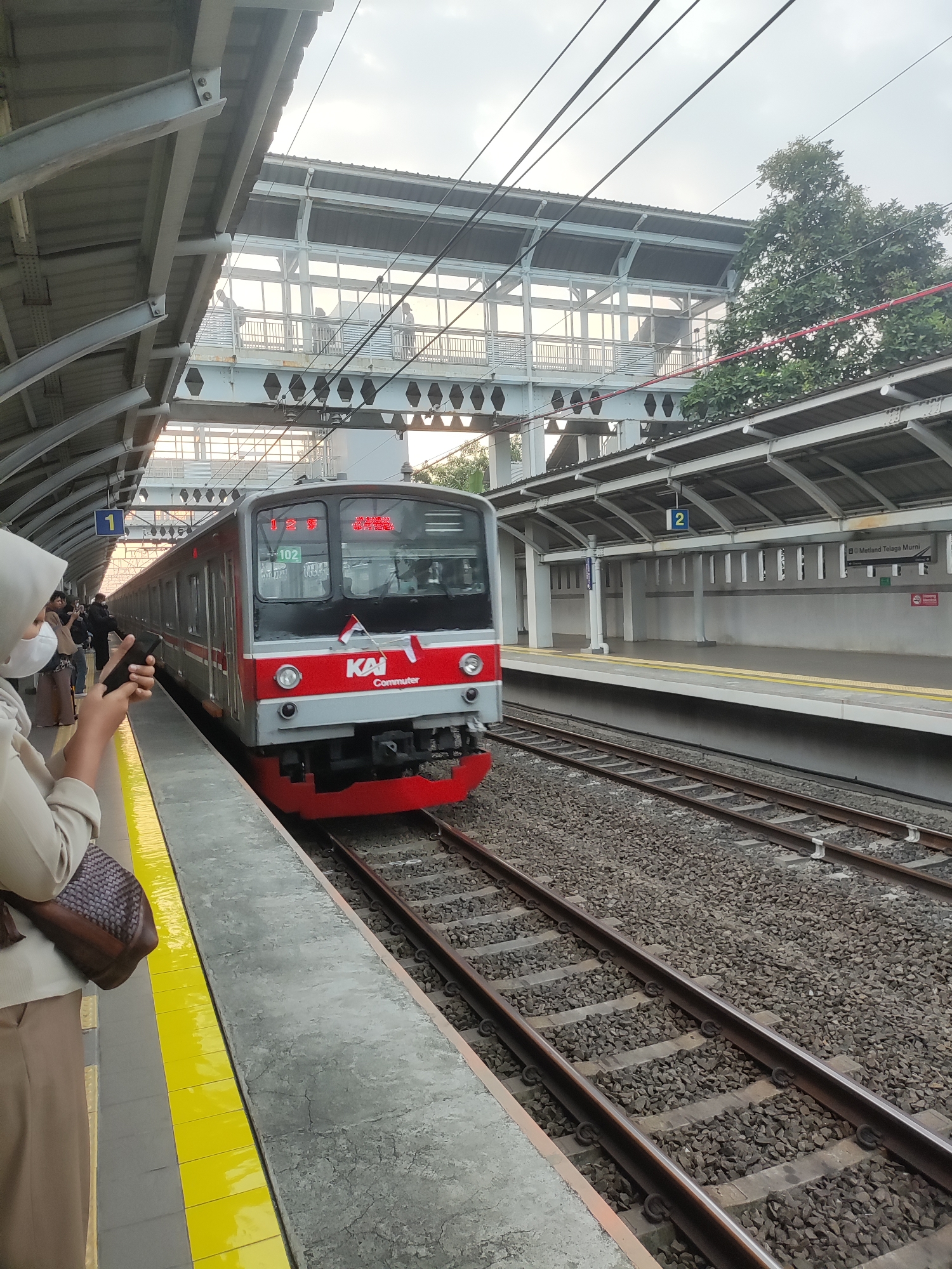 The image depicts a train at the Telagamurni station in Indonesia. A red and gray commuter train, numbered 102 and displaying "KAI Commuter", is stopped at the platform. The train has its headlights on and features a small Indonesian flag attached to its front. On the platform to the left, a person wearing a hijab and a light-colored jacket is holding a smartphone and a purse. Several other people are visible on the platform in the background, some appearing to be waiting for the train. The platform is lined with yellow safety markings. The station has a covered roof supported by metal beams, and overhead electrical lines are visible. To the right, another platform is present with a sign indicating "Metland Telaga Murni". The train tracks are made of ballast and sleepers. The sky appears overcast.