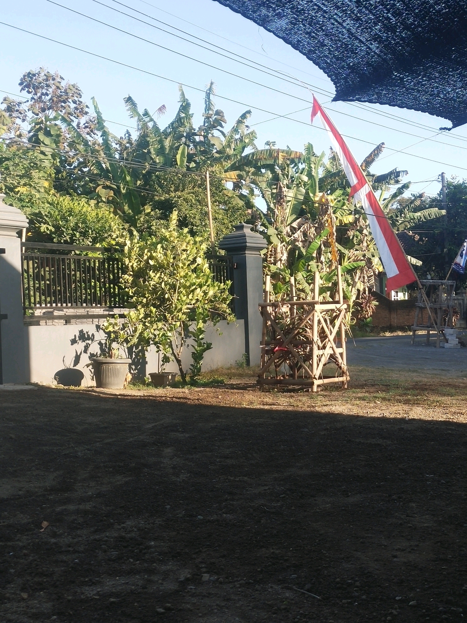 The image depicts a scene on a sunny day in Ambulu, Indonesia, likely in a residential or semi-rural area. In the foreground, a dark, rough-textured ground surface, possibly dirt or gravel, is visible. The middle ground features a grey wall with a black metal fence. To the left of the fence, a small tree in a dark pot casts a shadow. Further to the right, behind the fence, is a cluster of lush green trees, including banana trees. A red and white Indonesian flag is prominently displayed, tied to a wooden frame structure. In the background, there are more trees, a road, and a brick wall. Overhead, power lines crisscross the sky, partially obscured by a dark mesh netting in the top right corner. The overall impression is a bright, outdoor setting with elements of nature and human infrastructure.