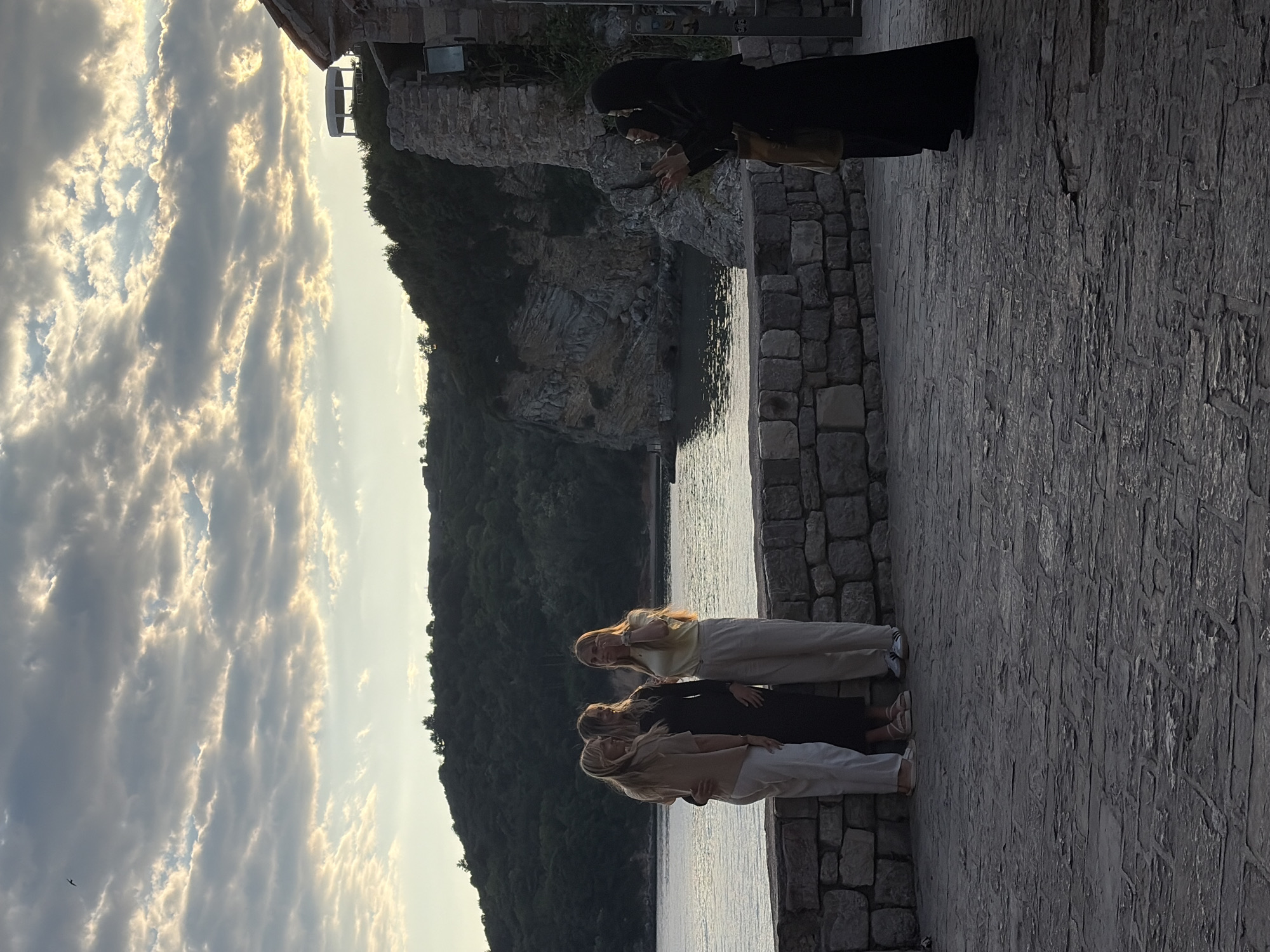 The image depicts a scene in Budva, Montenegro. In the foreground, three light-haired women stand on a stone-paved path next to the water. One woman is wearing a light yellow shirt, while another is wearing a black dress, and the third is wearing a beige shirt. Behind them is a lush, green hillside sloping down to the water. To the right, a woman dressed in a black abaya and hijab is taking a photograph with her phone. The background features a rocky cliff face topped with buildings and vegetation. The sky is filled with dramatic clouds and diffused sunlight.