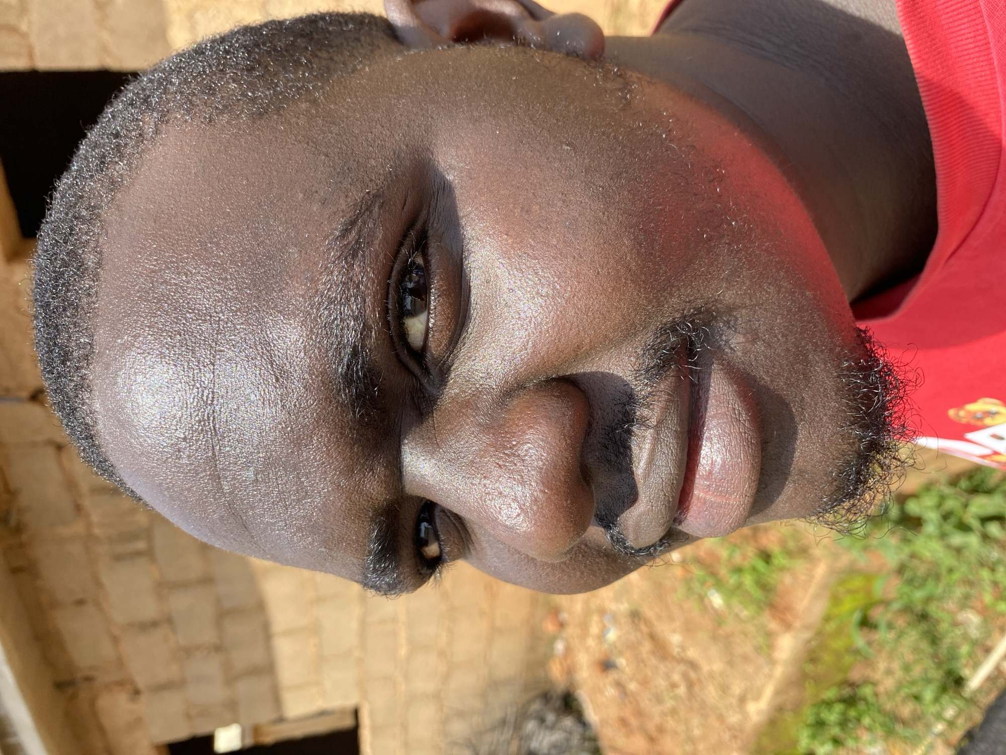 In Bukuru, Nigeria, a close-up, head-and-shoulders shot captures a man's face bathed in bright sunlight. He is looking towards the left side of the frame with a slight smirk. The man has dark skin, short, dark hair, and a goatee. His forehead appears shiny with sweat. In the background, a brick building with an open doorway is visible, along with some dry foliage and a hint of greenery.