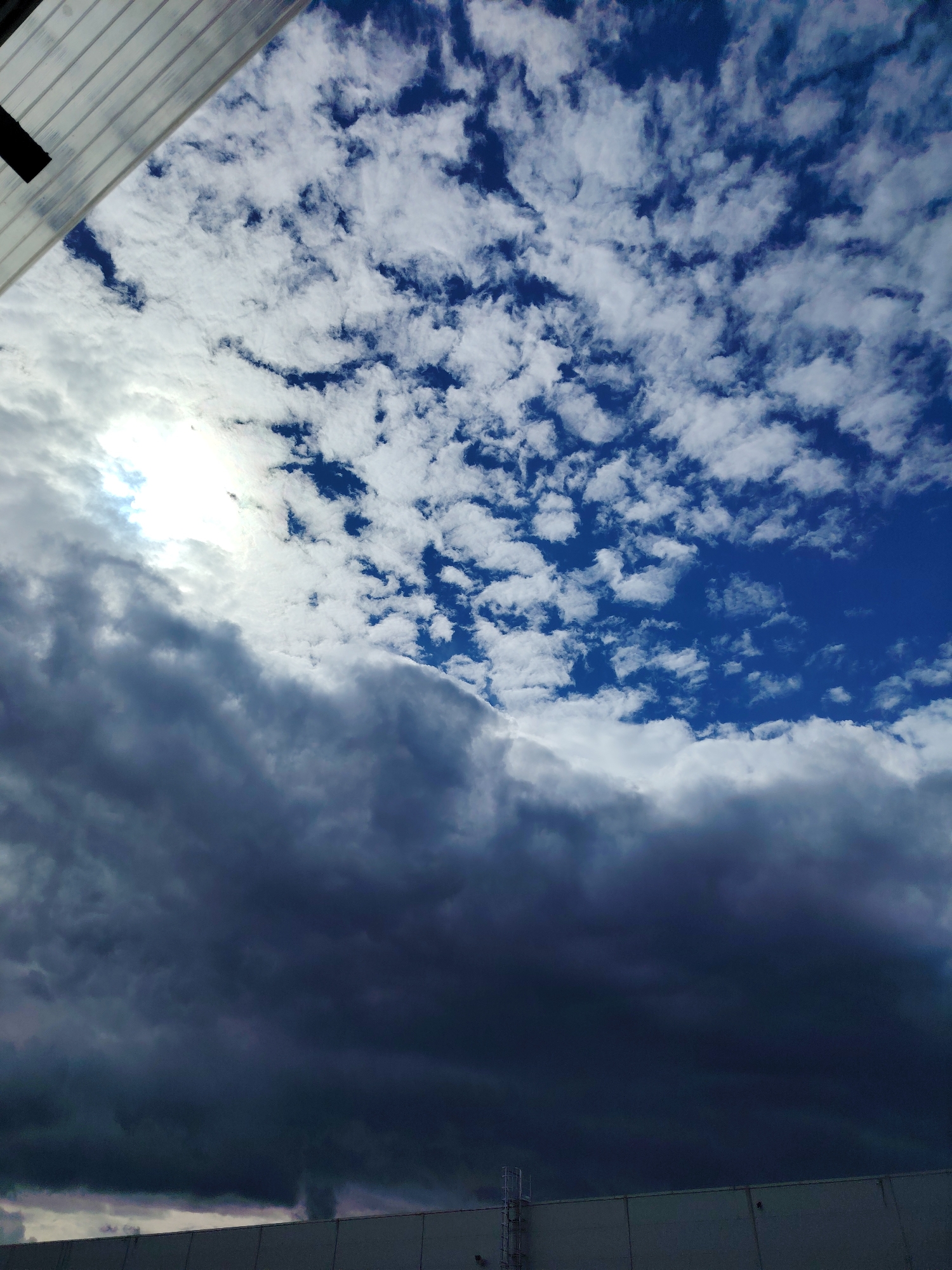 The image captures a dramatic sky over the city of Khorugvino, Russia. The upper portion of the frame is dominated by a bright blue sky filled with scattered white cumulus clouds. In contrast, the lower half of the image is obscured by dark, heavy storm clouds. The sun is partially visible through a break in the clouds on the left side, casting a bright light. In the foreground, a white, sloped structure, possibly part of a building or an overhang, is visible in the top left corner. At the bottom of the image, a long, light-colored wall stretches horizontally, with a metal ladder attached to it on the right side. The transition between the bright, clear sky and the ominous storm clouds creates a strong visual contrast.