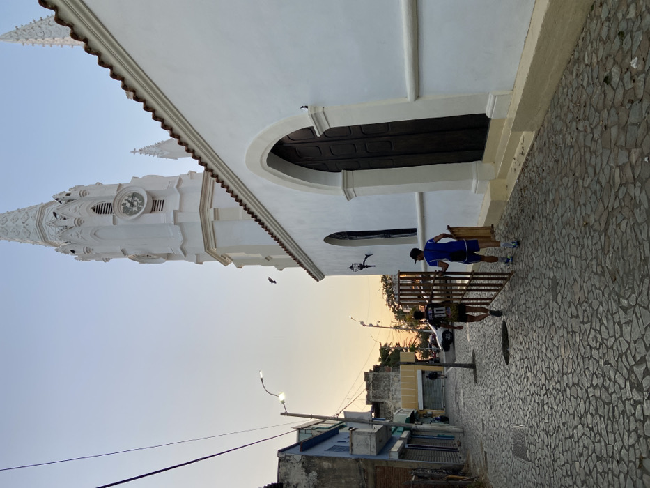 This image depicts a street scene in Juan Griego, Venezuela, during the early evening or late afternoon. On the right, the white Basílica Menor Nuestra Señora del Valle is prominently featured, characterized by its ornate, multi-spired steeple with a visible clock face, a terracotta-tiled roofline, and arched dark wooden doors on its facade. A black decorative wall lamp is affixed to the church's exterior.
The foreground and mid-ground are dominated by a cobblestone street, paved with irregular gray stones. Two young males are walking away from the camera along this street. The individual on the left wears a dark short-sleeved shirt with the number "10" visible on the back, and dark shorts. The individual on the right wears a blue short-sleeved shirt, blue shorts, and a baseball cap; he is actively holding and carrying a section of a dark metal gate with vertical bars in his left hand, and potentially a light-colored wooden board under his right arm.
To the left of the street, a series of single and two-story buildings line the roadway. These structures display varying architectural styles and include commercial establishments with closed metal roll-down security doors. One building on the far left is painted light blue. Overhead, multiple black power lines are strung between poles, one of which supports a modern LED streetlight. In the sky, which transitions from a light blue at the top to a warm orange-yellow near the horizon, a dark bird is visible in flight. In the far distance down the street, a white vehicle and another individual are faintly discernible.