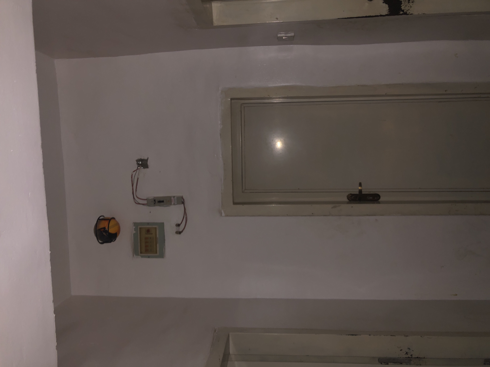 This is a full, eye-level shot of a hallway with a closed, light-colored door centered in the frame. To the left of the door is a white wall with a small, light-colored electrical panel mounted on it. Above the electrical panel is a round, orange and black object that appears to be a safety helmet. To the right of the electrical panel, a grey electrical box is mounted on the wall, with red wires extending from it. The hallway walls are painted a neutral, light color, and the ceiling is visible at the top of the frame, also painted white. The lighting is dim, casting a slight reflection on the door. The scene appears to be indoors, possibly in a residential or commercial building. The location is identified as Howayi, Nigeria.