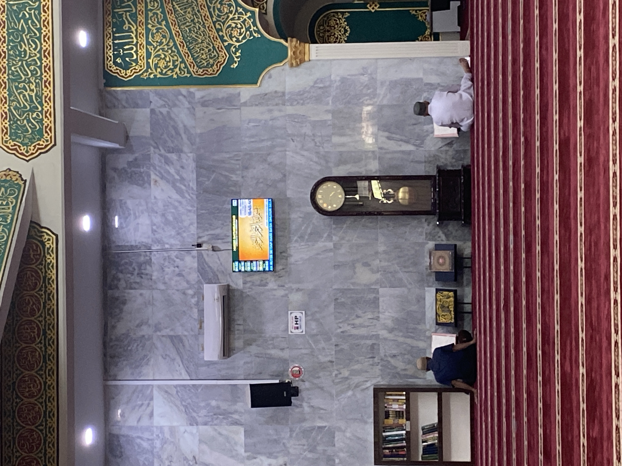 The image depicts the interior of a mosque, likely located in Lhokseumawe, Indonesia, given the prevalence of Islamic architecture and decor. The scene is dominated by a red carpet with gold stripes, which covers the floor and extends up the walls. The walls themselves are made of marble and are adorned with Arabic calligraphy in green and gold.
In the foreground, two men are seated on the carpet, facing a prayer niche. One man, wearing a blue shirt and a cap, is reading a book. The other man, in a white outfit and cap, is also reading.
In the background, a grandfather clock stands tall, and a television screen displays Arabic script. A bookshelf filled with books is visible to the left. An air conditioning unit is mounted high on the wall. The overall atmosphere is one of quiet devotion and reverence.