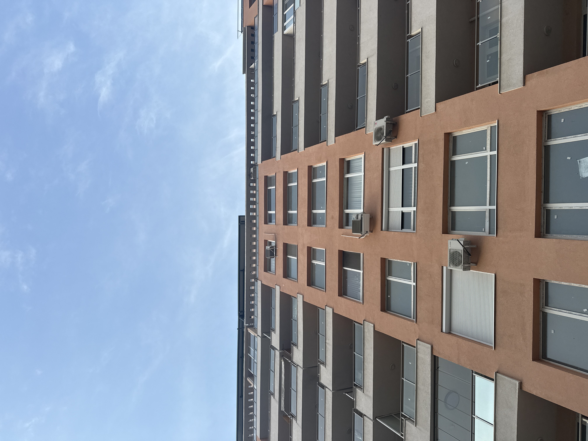 The image shows the exterior of a multi-story building in Plovdiv, Bulgaria. The building has a façade with alternating sections of terracotta-colored stucco and light gray concrete balconies. Multiple windows with white frames are visible, some with curtains or blinds. Several air conditioning units are mounted on the terracotta sections of the building. The sky is visible to the side of the building, showing light blue with sparse clouds. The image is taken from a low angle, looking upwards at the building.