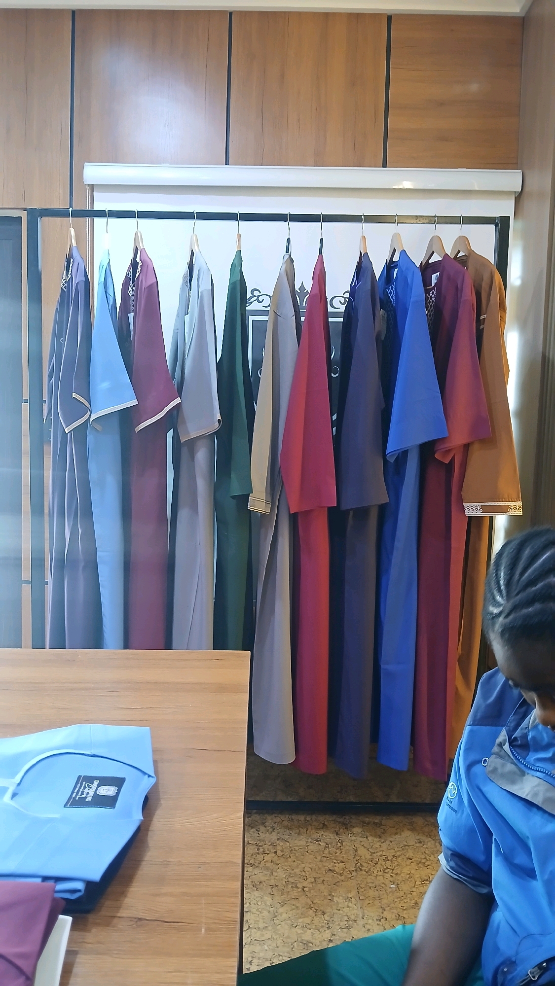 The image depicts an indoor setting, likely a clothing store or workshop in Gura Top, Nigeria. A rack of immaculately hung garments, predominantly in muted earth tones and some vibrant jewel colors like maroon and royal blue, dominates the background. These appear to be traditional long robes or professional attire, with some featuring decorative trim. The garments are hung on identical wooden hangers, creating a neat and organized display.
In the foreground, a wooden table is visible, with a light blue garment folded on its surface. A black label with text and a logo is attached to the collar of this garment. Part of a person, wearing a blue and grey jacket, is visible on the right side of the frame, partially obscuring the clothing rack. Their hair is styled in braids. The floor appears to be covered in a textured material, possibly cork or a similar composite, with a speckled pattern. The overall lighting suggests an interior environment, possibly with ambient light from windows or overhead fixtures, creating soft shadows and highlighting the textures of the fabrics and wood. The environment appears clean and orderly, suggesting a professional or retail space.