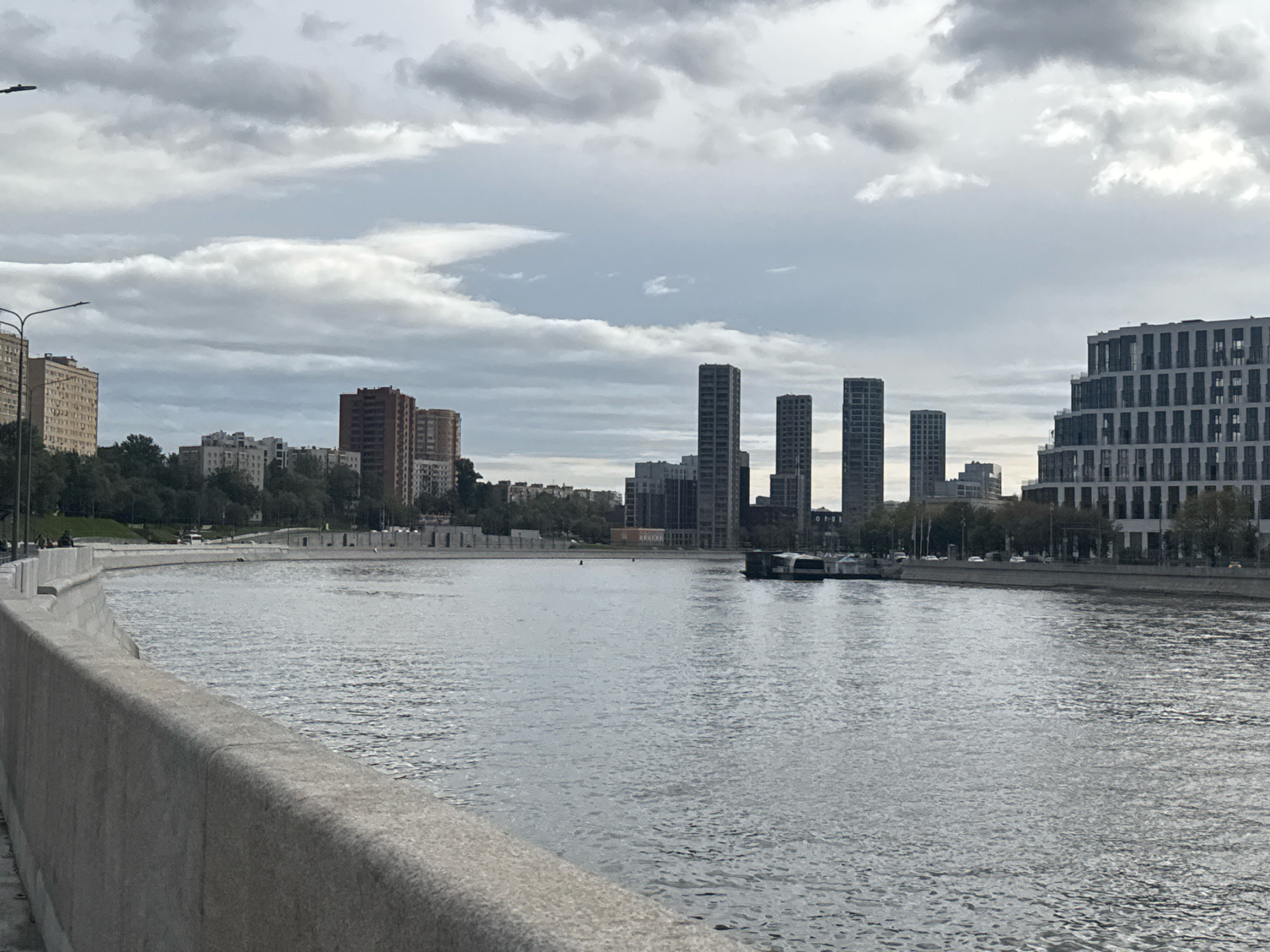The image shows a wide river in Moscow, Russia, on a cloudy day. A concrete embankment lines the left side of the river, with a pedestrian walkway visible behind it. On the far side of the river, a cityscape unfolds, featuring various buildings ranging from mid-rise residential complexes to modern high-rise towers. Notably, several tall, narrow skyscrapers are clustered together, and a larger, curved building with many windows is on the right. Trees and a road with cars are also visible along the embankment. A dark, rectangular boat or barge is docked on the water in the middle distance. The sky is filled with heavy, grey clouds, creating a diffused light over the scene. There are no discernible people or animals in the foreground or midground.