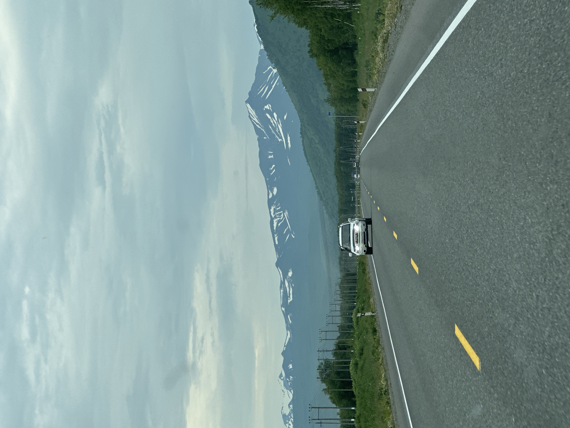 An eye-level, point-of-view shot depicts a car driving on a two-lane asphalt road, which stretches into the distance. The road is marked with solid white lines on the right and dashed yellow lines on the left, indicating traffic flow. The sky above is overcast with light gray and white clouds. In the middle ground, on either side of the road, there is a grassy shoulder lined with utility poles. Further back, a dense line of green trees forms a backdrop, beyond which lie majestic mountains with snow-capped peaks. A white SUV is visible on the road, driving towards the viewer. There are no people visible in the image. The scene appears to be set in a rural or mountainous area, possibly in the vicinity of Кеткино, Russia, given the presence of utility poles and the natural landscape.