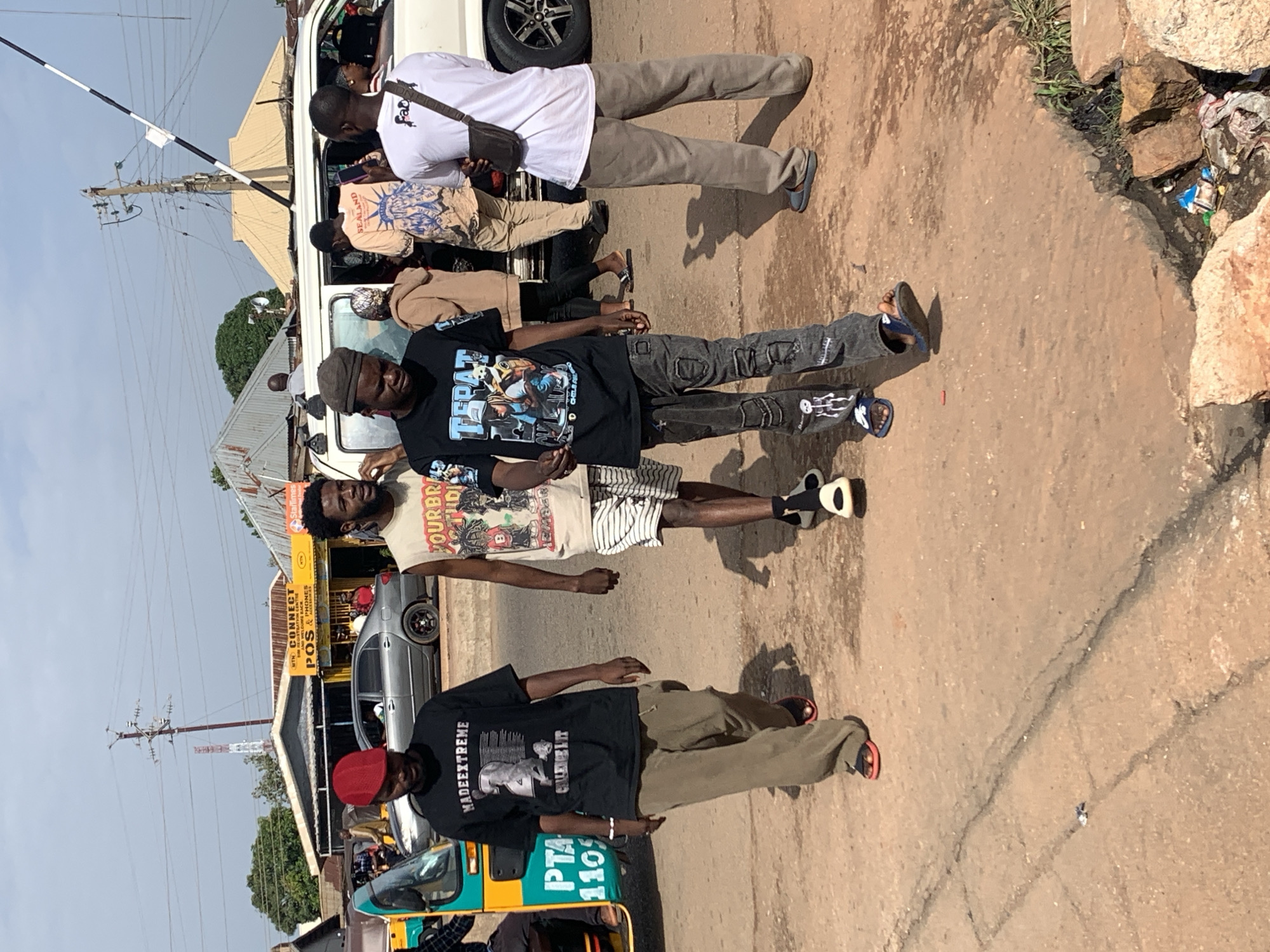A group of four young men are walking on a paved road in what appears to be a commercial area in Gura Top, Nigeria. The man on the far left is wearing a red cap, a black t-shirt with graphics, and khaki pants. Next to him is a man in a light-colored sleeveless shirt and striped shorts. The third man from the left is wearing a black t-shirt with graphics and dark distressed jeans. The man on the far right is wearing a white t-shirt with a dark shoulder bag and grey sweatpants. In the background, there are buildings, vehicles including a white van and a tricycle, and utility poles with wires. The ground is paved with some cracks and debris.