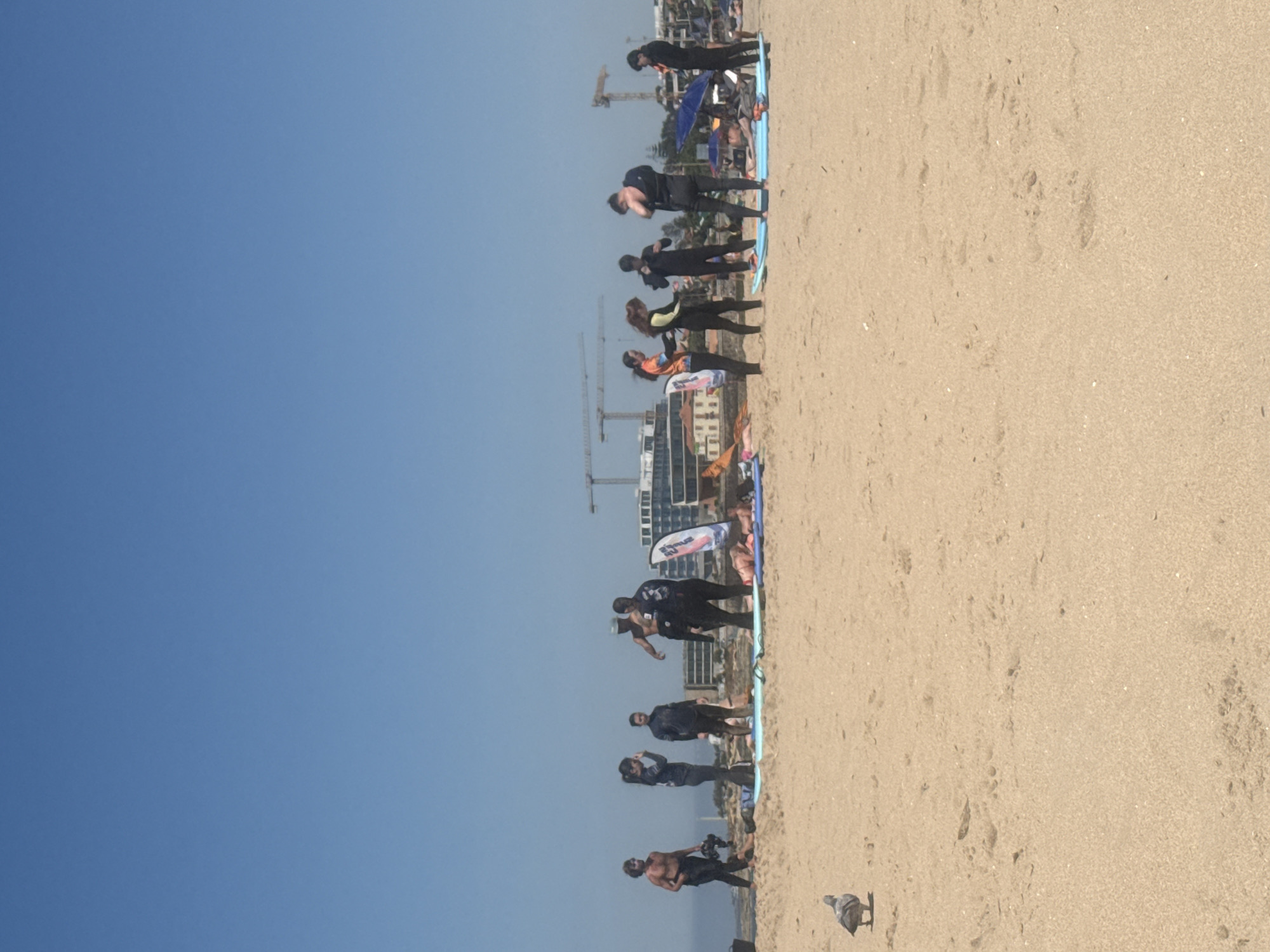 The image captures a sunny day at Guincho Beach in Cascais, Portugal, with a group of people in wetsuits standing on the sand. A pigeon is visible in the foreground. In the background, there are buildings and construction cranes, indicating ongoing development. The beach is wide and sandy, with the ocean stretching out to the horizon under a clear blue sky. The people appear to be engaged in some sort of activity, possibly a surfing lesson or practice session, with surfboards laid out on the sand. Some individuals are interacting with each other, while others are focused on their own movements. The scene conveys a sense of outdoor recreation and a vibrant coastal atmosphere.