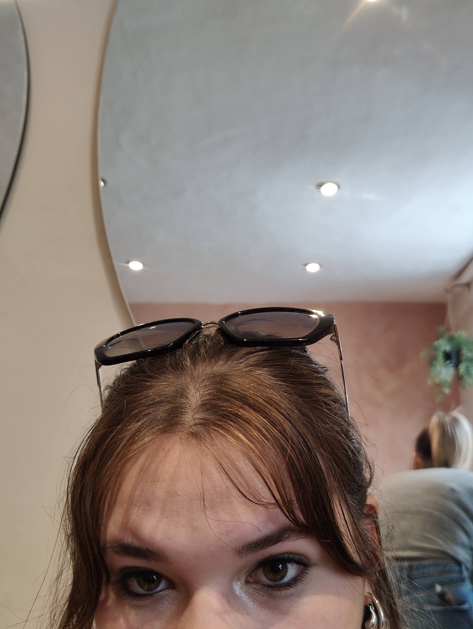 The image is a close-up shot of a person's face from a low angle, with sunglasses resting on their head. The person has light brown hair with bangs, brown eyes, and fair skin. They are wearing silver hoop earrings and eyeliner. The background includes a mirror on the left, a ceiling with recessed lighting, a wall painted a light peach color, and a blurred image of another person with blonde hair and a gray shirt. There is also a plant visible in the background. It is not possible to determine whether the location is in Ipswich, United Kingdom, from the image alone.