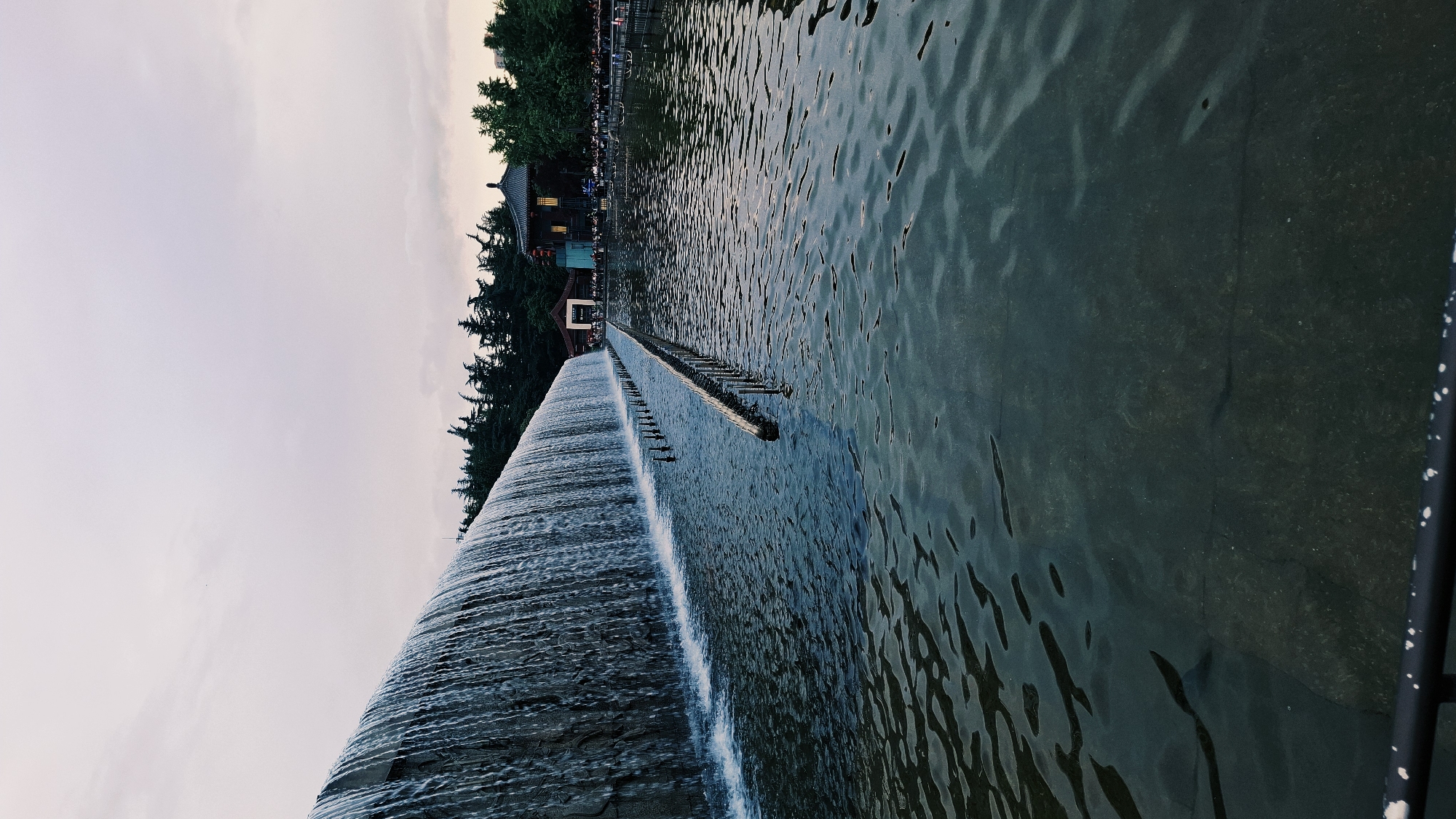 A low-angle, wide shot depicts a man-made waterfall in Yanta District, China. The water cascades over a concrete structure, creating a sheet of water. Above the waterfall, the water's surface ripples. A row of metal posts extends along the top of the dam. Beyond, trees line the water's edge, interspersed with buildings. On the left, the sky appears overcast. A black metal railing is on the lower right.