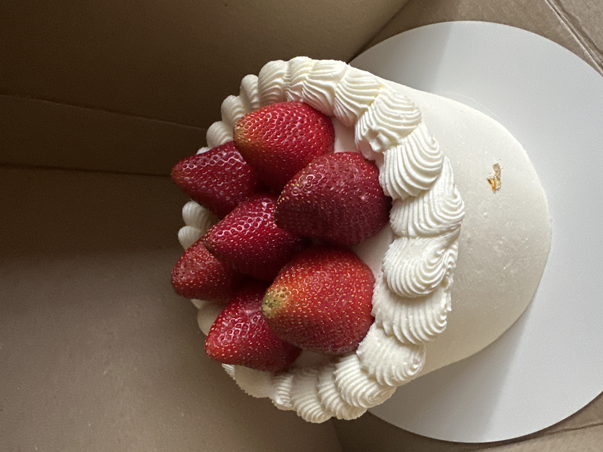 A top-down shot shows a cake with white frosting and strawberries in a box. The cake has a white frosted top, surrounded by piped white frosting. The strawberries are a deep red color. The box is brown. A small piece of gold flake decorates the frosting. There is no indication that this image was taken in Ternopil, Ukraine.
