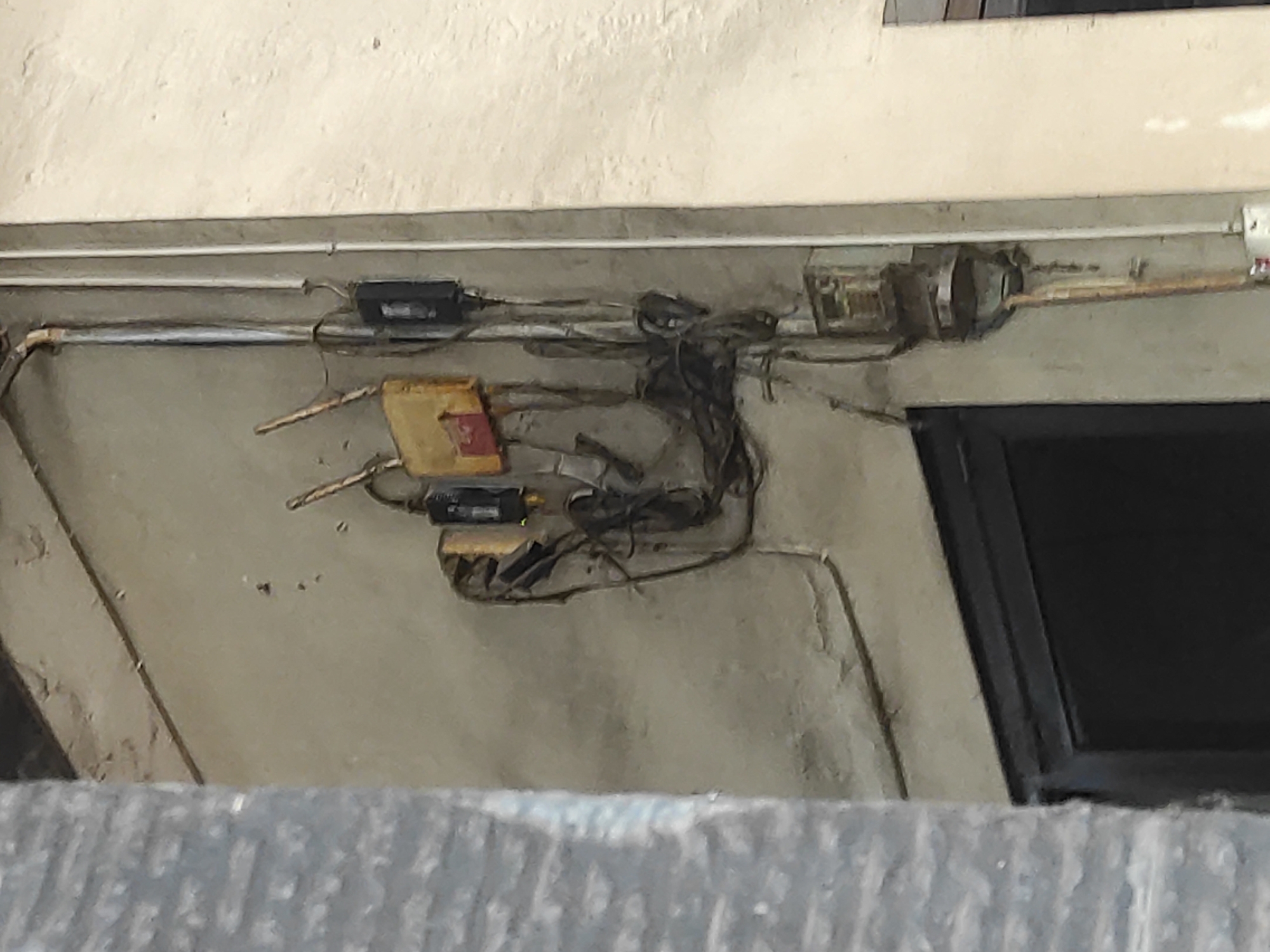 The image depicts an outdoor scene on a wall in Lhokseumawe, Indonesia. The wall is made of concrete and appears aged, with some signs of wear and tear. Attached to the wall are various electrical components. There are several black electrical boxes, some with visible wires and conduits. One box has a yellow cover with a red insert. A metal pipe runs vertically along the wall. Below these components, a dark-colored door or window frame is visible. To the right of the electrical fittings, a lighter-colored wall section is present. The overall impression is that of an electrical junction or distribution point on the exterior of a building.