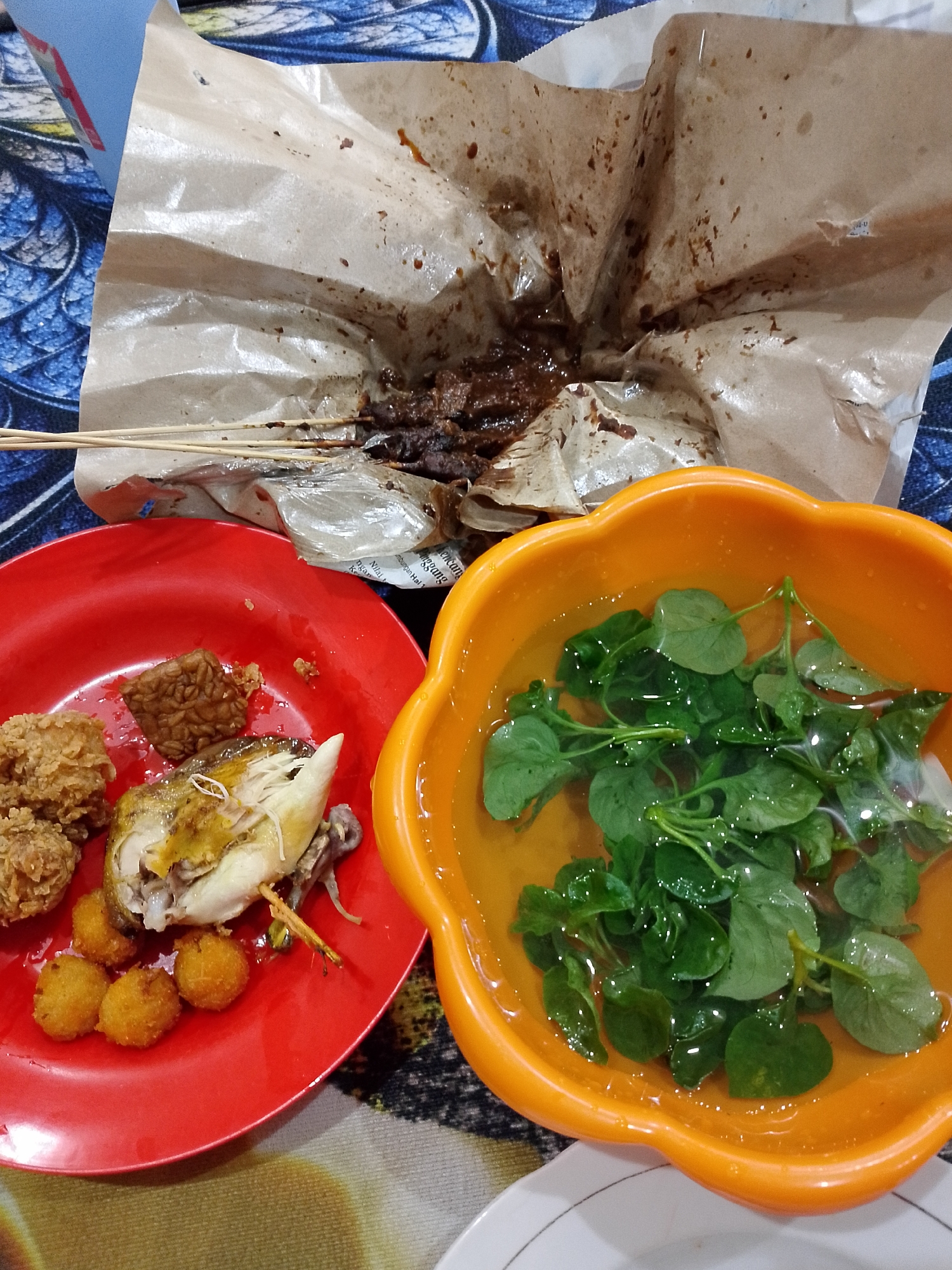 In Balikpapan, Indonesia, a close-up, eye-level shot captures a meal laid out on a patterned tablecloth. The scene is dominated by a red plate holding pieces of fried chicken, battered balls, tempeh, and a portion of a roasted chicken. Adjacent to the red plate is an orange bowl filled with a clear liquid and fresh green leaves, likely a side dish or soup. In the background, a piece of brown paper wrapper, stained with dark sauce and holding several skewers of meat, suggests a takeaway or shared meal. The lighting is moderate, highlighting the textures and colors of the food. No individuals are visible in the frame.