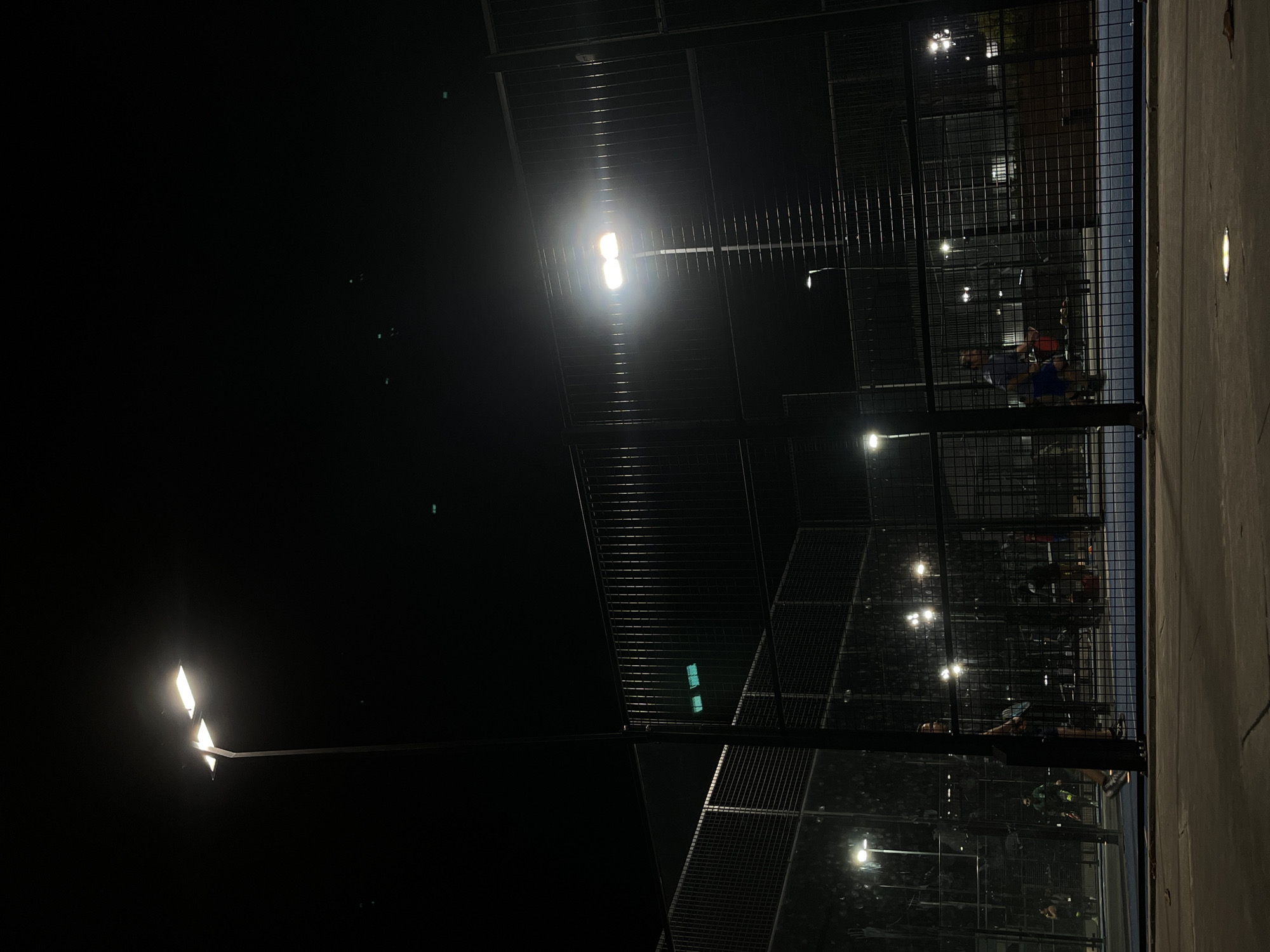The image captures an outdoor sports court, likely for padel or tennis, illuminated at night. The scene is dominated by the deep black of the night sky, contrasting sharply with the bright, artificial lighting.
The court is enclosed by tall, dark wire mesh fences that create a grid-like pattern across the middle and right portions of the image. Several rectangular floodlights are mounted on poles, casting a powerful, white glow onto the court. One prominent light source is centered in the upper half, while another is on a pole extending from the left side. Additional, less distinct lights can be seen through the mesh in the background.
Several people are visible within the court enclosure, engaged in what appears to be a game or practice session. In the foreground, viewed through the mesh, two individuals are somewhat clearer; one wears a blue top and the other a darker shirt, both seemingly focused on the activity. Other figures are visible deeper within the court, partially obscured by the fencing and shadows, but their presence indicates ongoing play.
On the right side of the image, a solid, light-colored wall stands vertically, with a small, embedded light fixture visible on its surface. The court surface itself appears dark. The time of day is clearly night, as evidenced by the profound darkness and the reliance on the numerous, bright floodlights to make the activity possible. No specific weather conditions are discernible, and there is no visible text in the image.